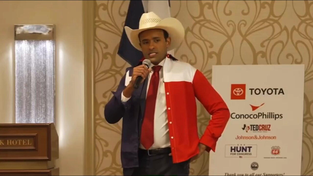 A picture of a grimacing Vivek Ramaswamy in a large white cowboy hat and a Texas-themed coat making a speech while standing in front of a bunch of corporate logos at what looks like a hotel event space.