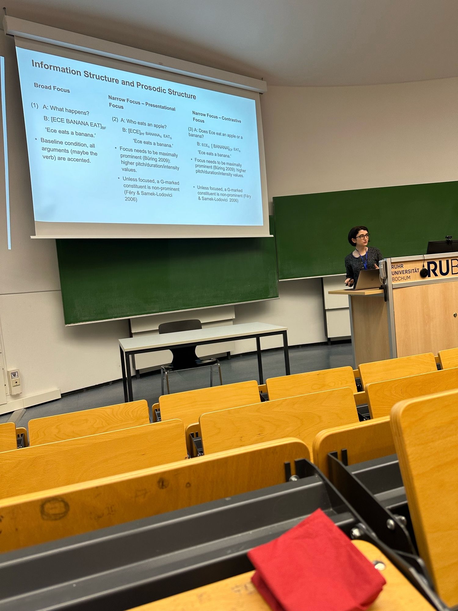 At a classroom, a woman with glasses standing in front of a presentation. The presentation shows information structure and prosody in spoken languages.