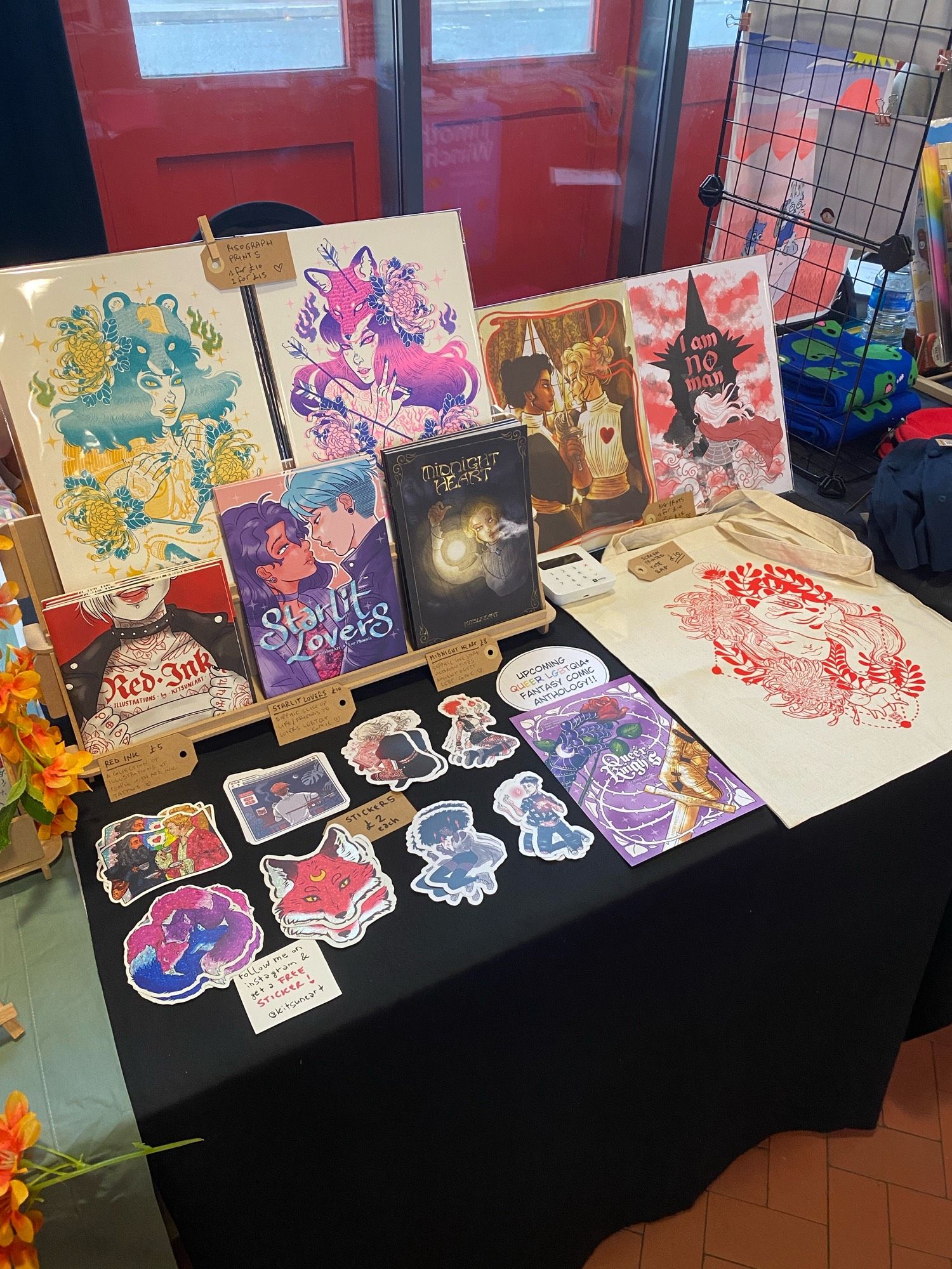 Close up on the table display showcasing Starlit Lovers comic, Midnight Heart comic and Red ink zine, plus various stickers, riso graph prints and tote bag