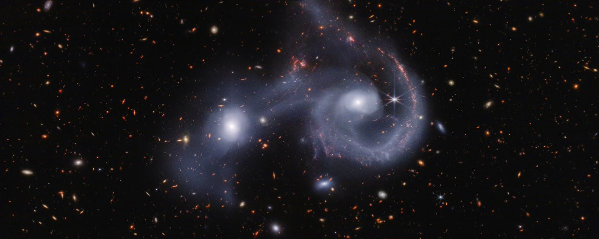 A pair of interacting galaxies. The larger of the two galaxies is slightly right of center, and composed of a hazy, bright, white center and a ring of gaseous filaments, which are different shades of white with some pink higlights. Toward the bottom left and bottom right of the ring are filaments of gas spiraling inward toward the core. At the top left of the ring is a noticeable gap, bordered by two pinkish orange pockets of dust and gas. The smaller galaxy to its left is made of hazy white gas and dust, which becomes more diffuse farther away from its center. To this galaxy’s bottom left, there is a smaller, more diffuse gas cloud that wafts outward toward the edges. Many red, orange, and white background galaxies are spread throughout, with some hazier in composition and others having more defined spiral patterns.