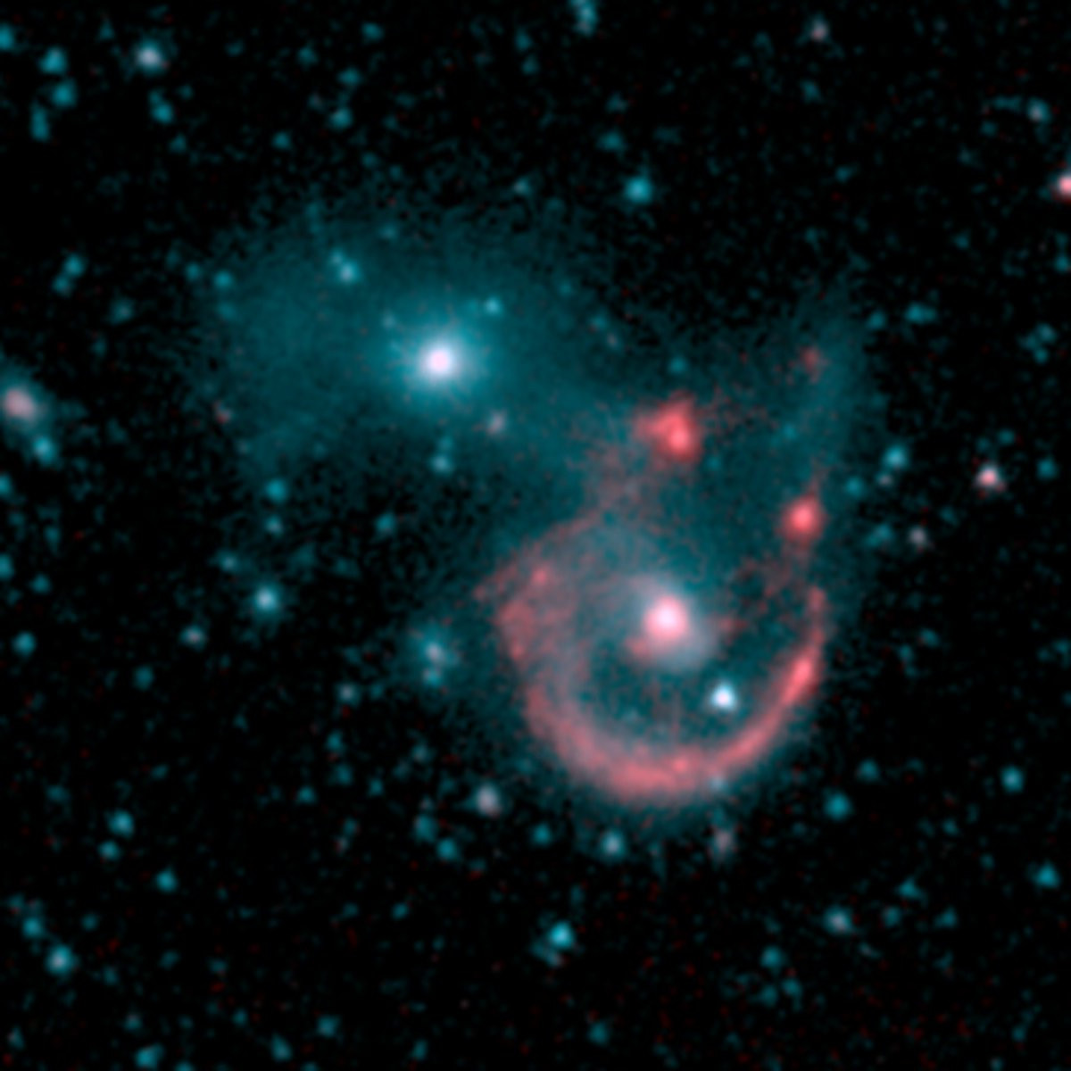 Spitzer’s infrared image is more diffused than either Hubble or Webb. Just right of center is a bright red dot, which is the center of the spiral galaxy. It is surrounded by a reddish ring, which are the galaxy’s arms.  The gap (here at top right) is bordered by two reddish pockets of dust and gas.  The smaller galaxy is at top left. It’s a bright blue dot surrounded by light blue haze. Background galaxies and stars are hazy blue dots. 