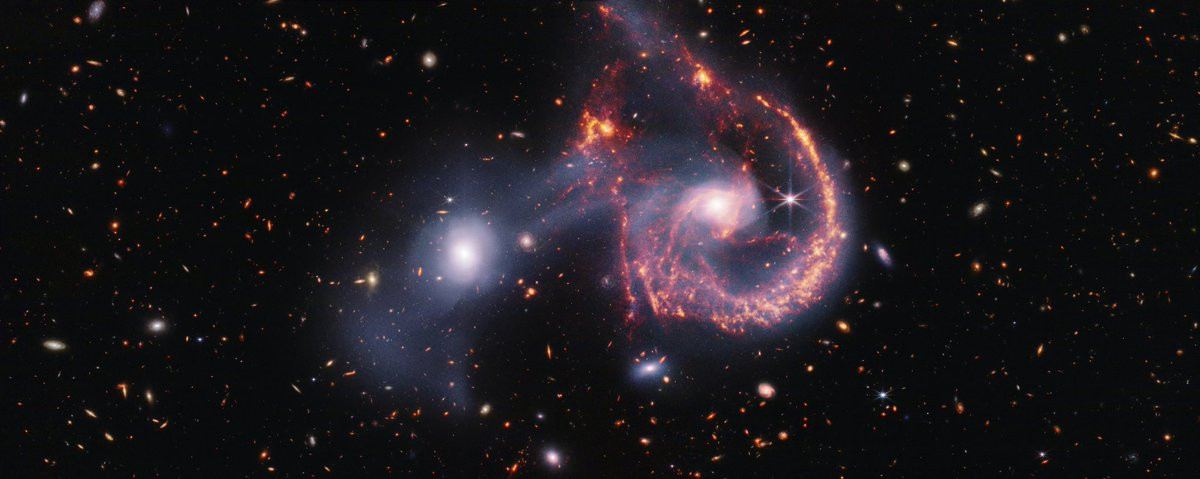 A pair of interacting galaxies. The larger of the two galaxies is slightly right of center, and composed of a hazy, bright, white center and a ring of gaseous filaments, which are different shades of red and orange. Toward the bottom left and bottom right of the ring are filaments of gas spiraling inward toward the core. At the top left of the ring is a noticeable gap, bordered by two large, orange pockets of dust and gas. The smaller galaxy to its left is made of hazy white gas and dust, which becomes more diffuse farther away from its center. To this galaxy’s bottom left, there is a smaller, more diffuse gas cloud that wafts outward toward the edges. Many red, orange, and white background galaxies are spread throughout, with some hazier in composition and others having more defined spiral patterns.