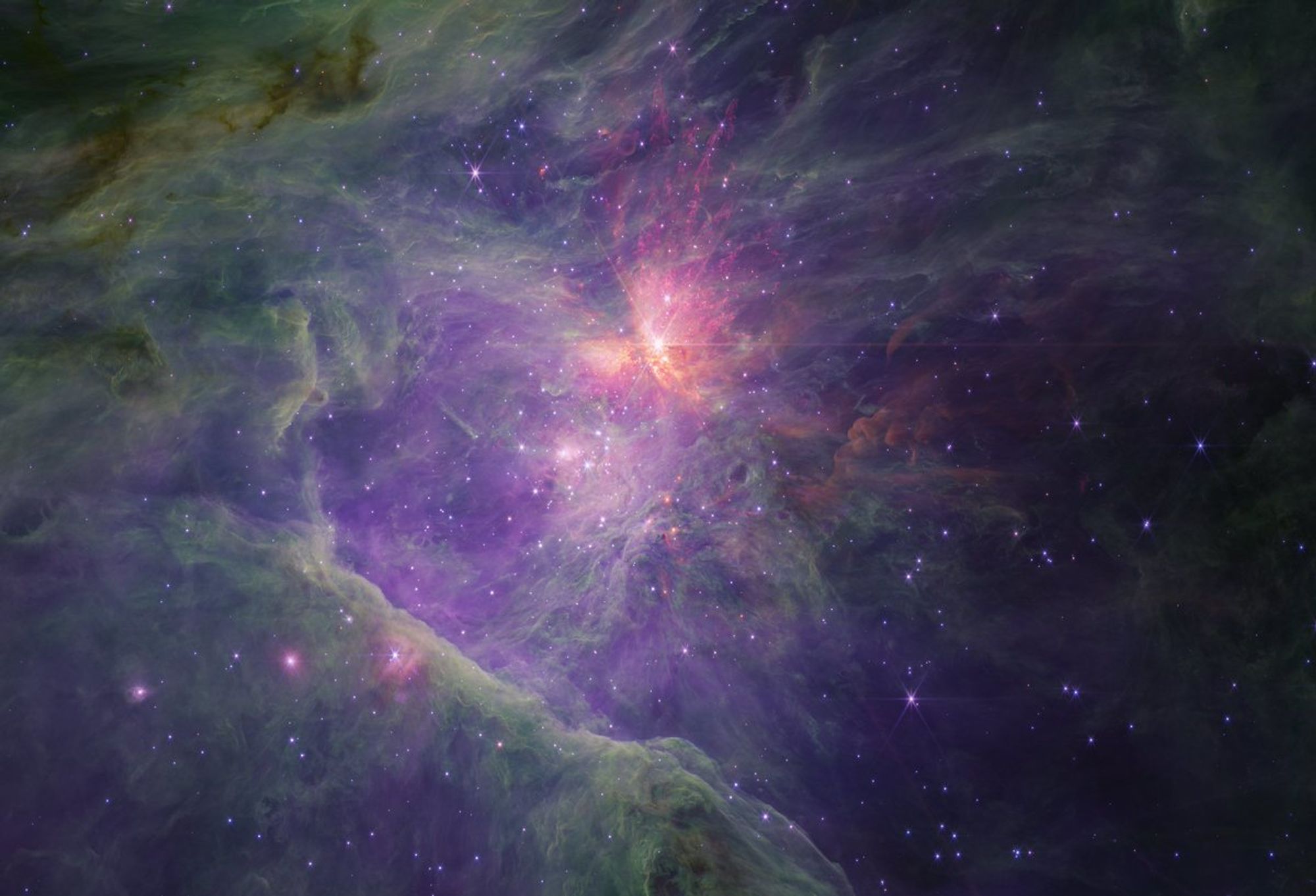 An image of a young star-forming region filled with with wispy purple, green, and red nebulosity. The purple ionized gas is seen mostly towards the center, with browns, greens, and reds behind, while the periphery is mostly bright green and darker brown to the left. There is a large spray of yellow, orange, red, and purple towards the top center, and the nebula fades to near black to the right. There are thousands of stars sprinkled across the field, concentrated towards the center, but they generally appear fainter at longer wavelengths, with some exceptions. The