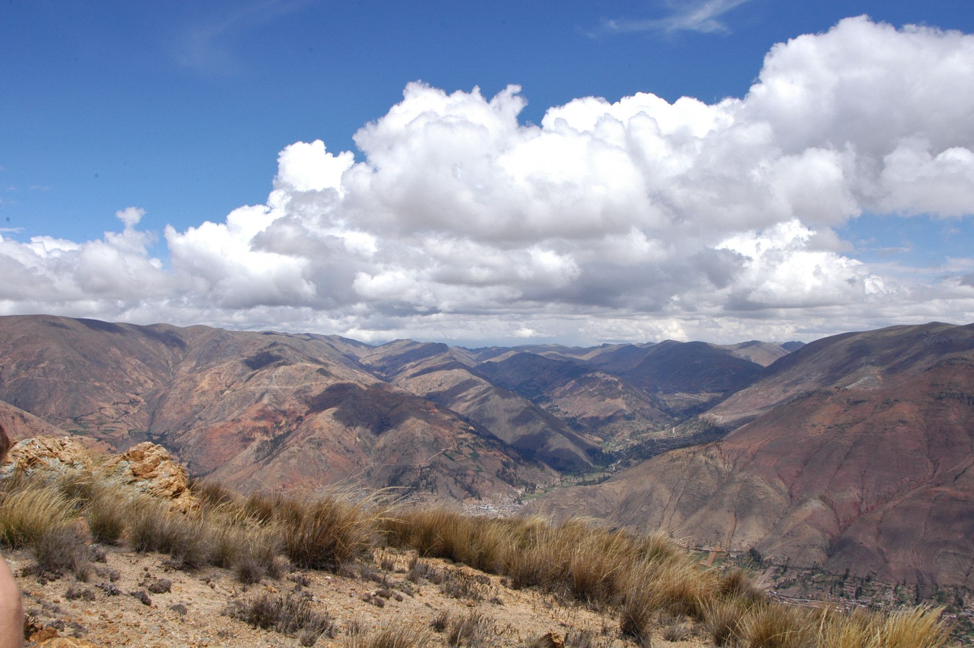 Au-dessus de 3000m, la vallée de Tarma sous un ciel bleu et de nuage blanc, elle s'étend entre zone ensoleillée et sous l'ombre des nuage, en 1er plan, quelques plantes rases sur un sol en légère descente..