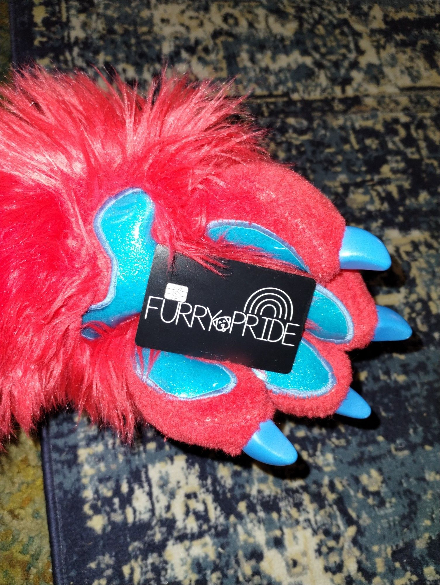 A red coyote paw with blue sparkling paw pads is holding a black cash app card that has a custom design, which is the words Furry Pride, with a dog in between the words and a rainbow above them.