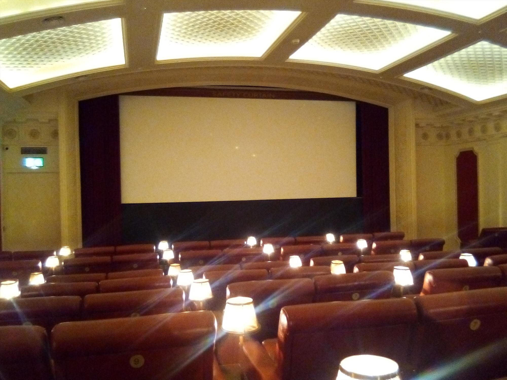 Inside Grosvenor Picture House, Glasgow; a small empty cinema room, with an arched cream colour ceiling and walls, large flat panel ceiling lights, red seats, with seat adjacent lamps, and an empty cinema screen.