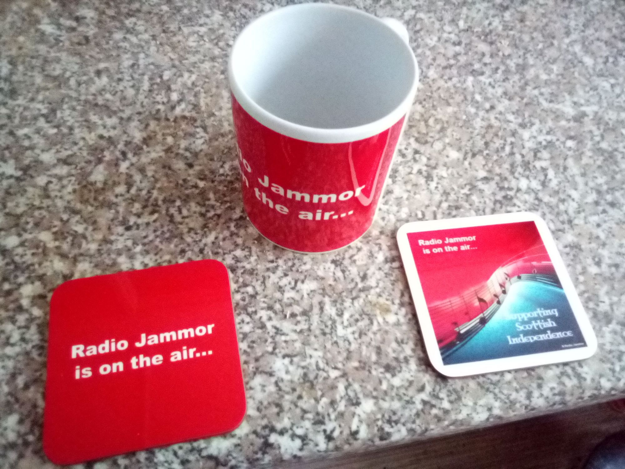 Radio Jammor is on the air... (RJIOTA) mug, plus two coasters, one with a plain RJIOTA design, the other with a red/blue split image with RJIOTA on the red, with music notes weaving through the image to separate red from blue and "Supporting Scottish Independence" text on the blue.