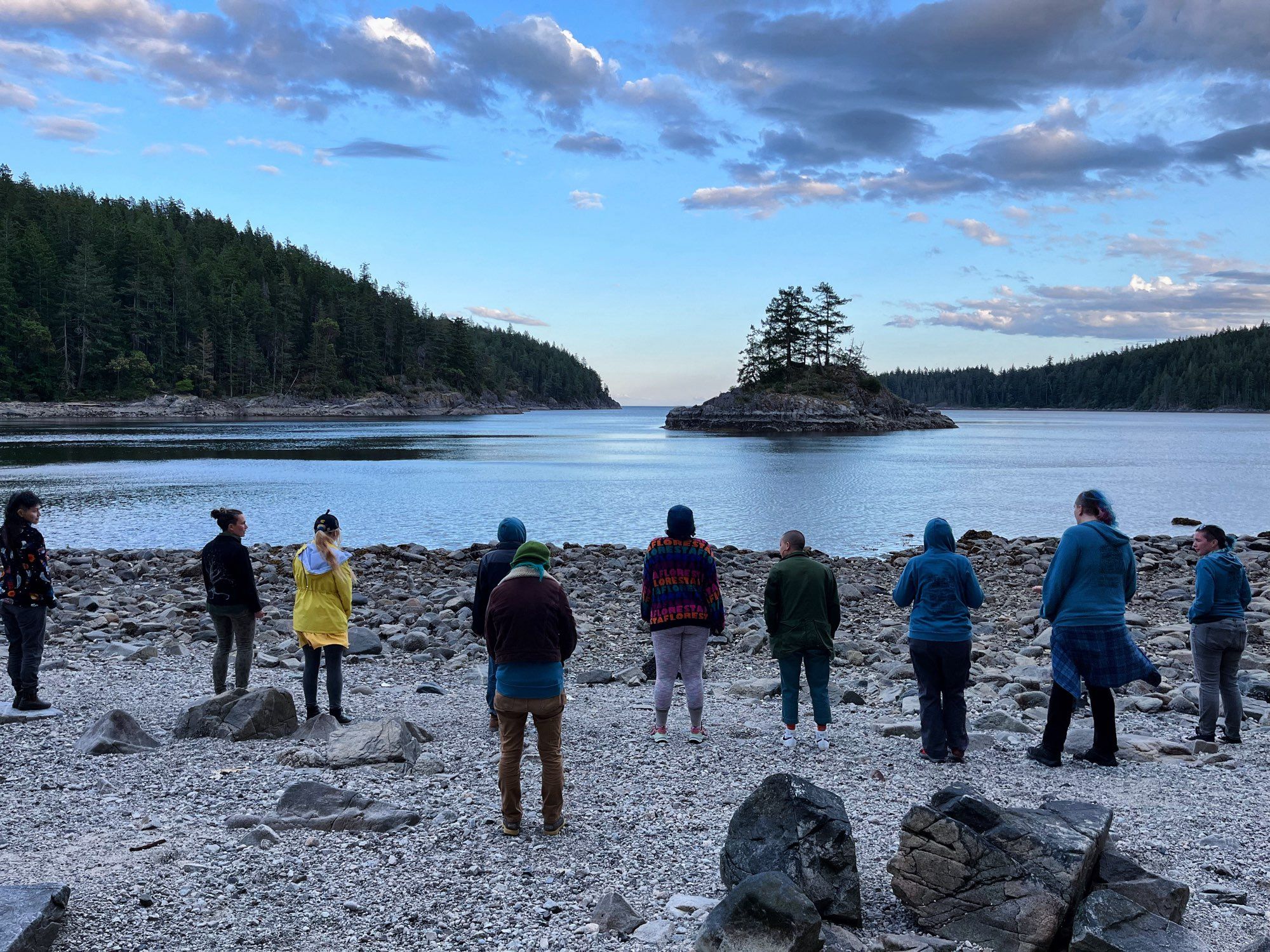 Nine retreaters stand in a line on a rocky beach, looking out over the water toward wooded shores and a small island in the bay.