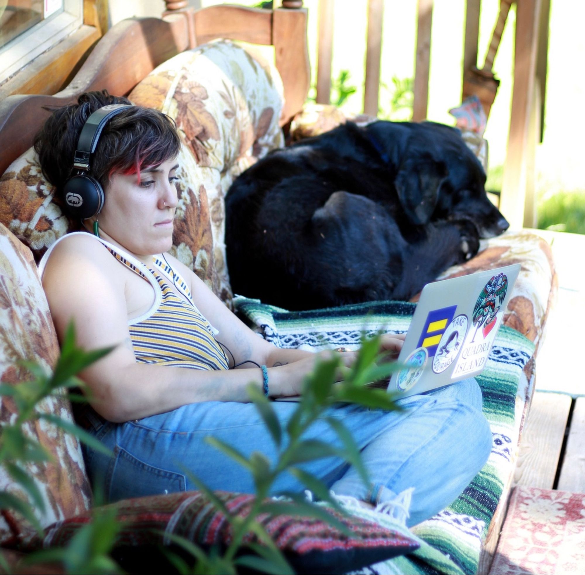 A retreater works on a laptop on an outdoor couch shared by a black labrador dog.