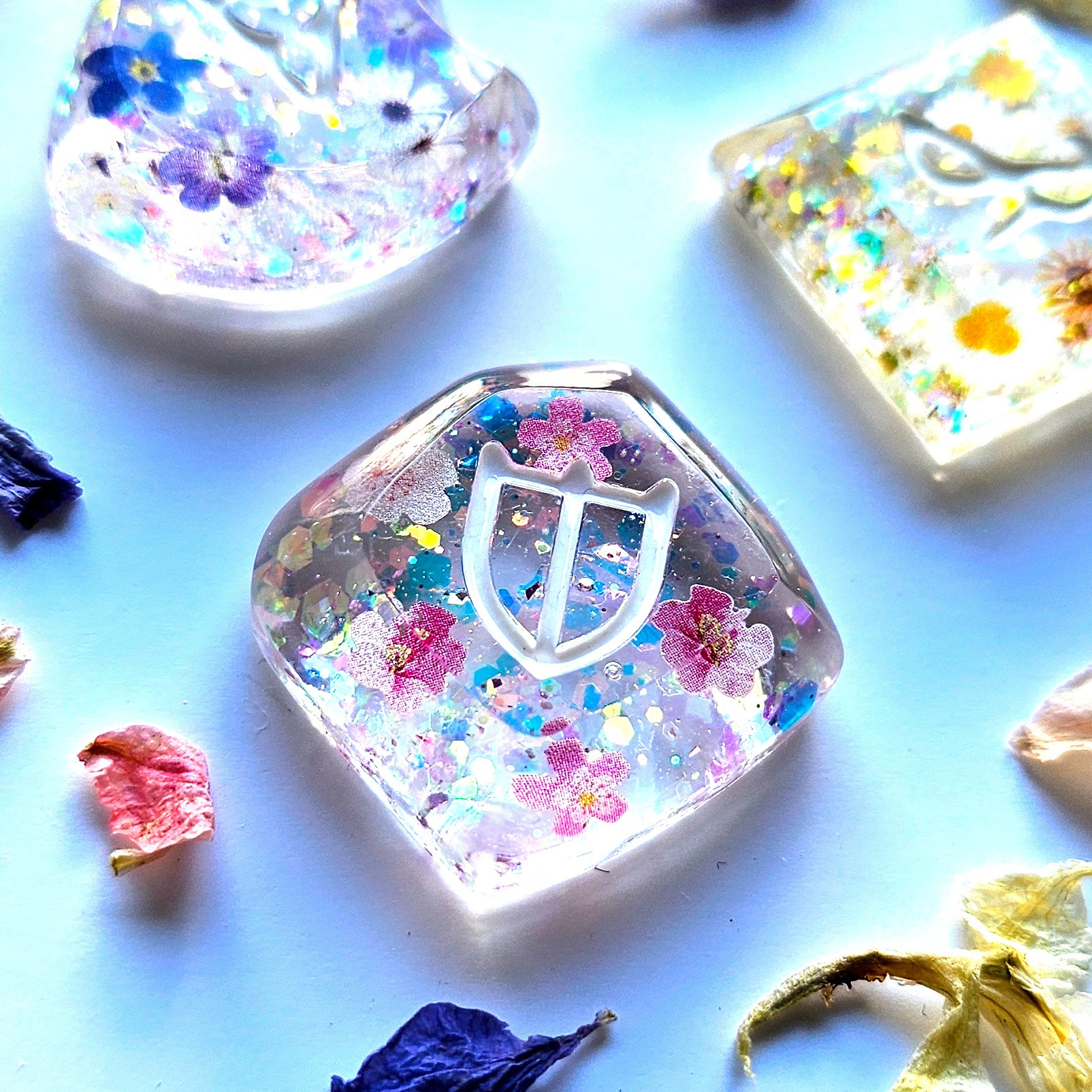 A zoom photo of a paladin resin job stone inspired from the game final fantasy 14.
It's transparent with gold and blue glitter, as well some pink flower elements in it. Around it are dried flowers petals that are yellow, pink and purple.