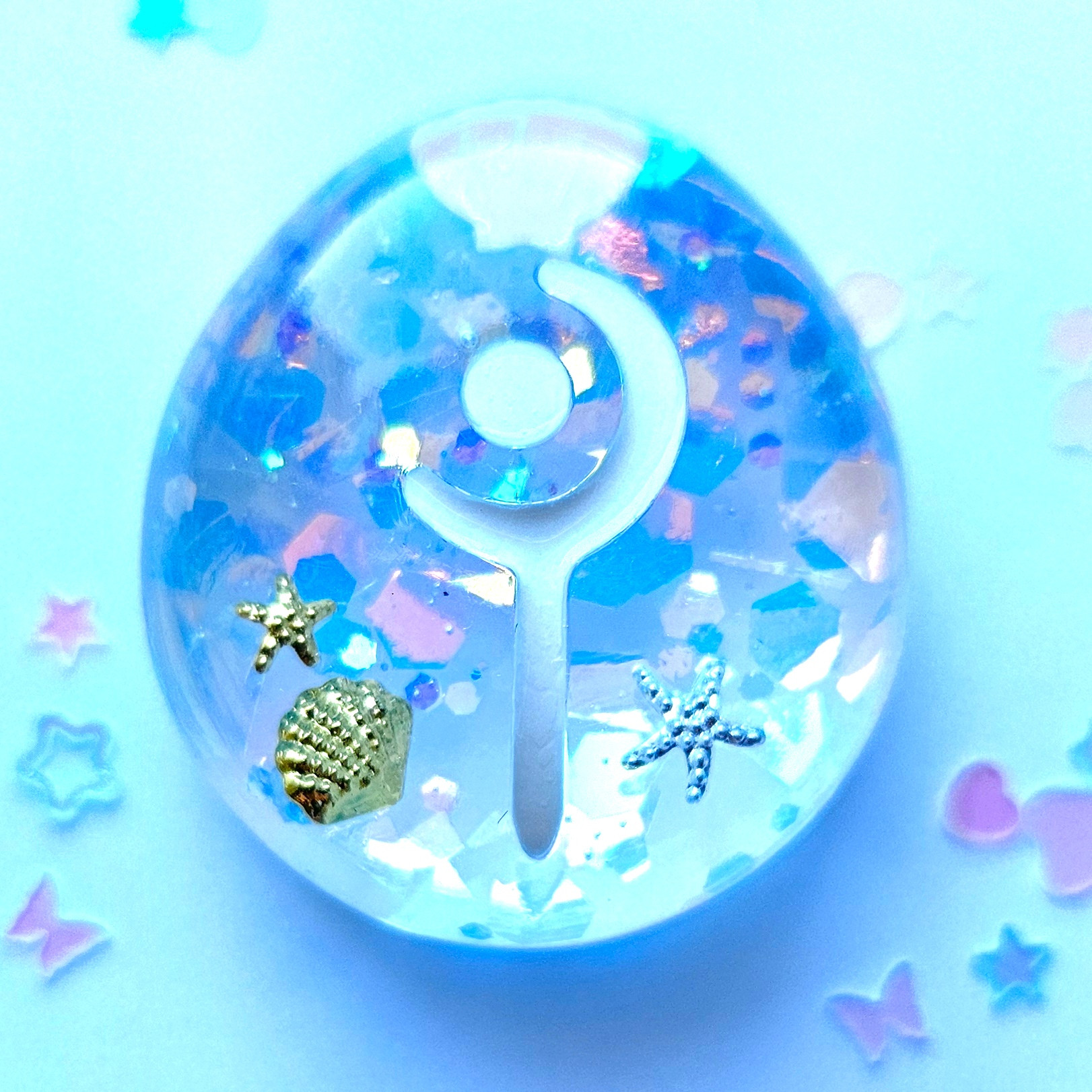 A zoom photo of a white mage resin job stone inspired from the game final fantasy 14.
It's transparent with blue and iridescent glitter, as well some gold and silver sea star and seashell elements in it. Around it are pastel pink and blue glitter with heart and star shapes.
