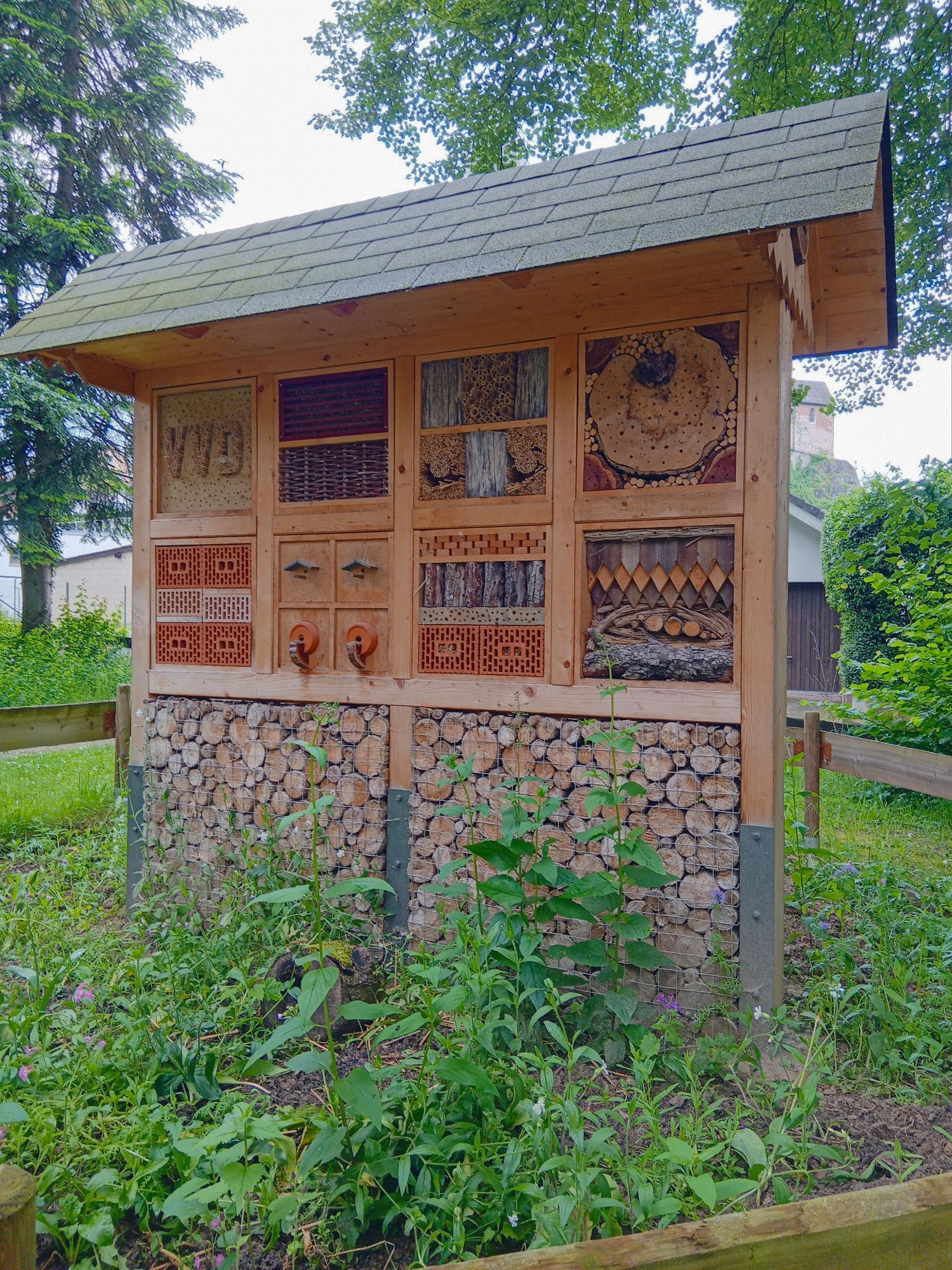 Ein aus verschiedenen Brettern gezimmerter Holzstand mit einem großen Giebeldach. Der Holzstand ist in acht quadratische Segmente unterteilt, die wiederum quadratisch aufgeteilt und farbig angestrichen sind. Diese Segmente beherbergen Insektenvölker. Dieses Insektenhotel steht eingezäunt inmitten von wilden Gräsern und Stauden.