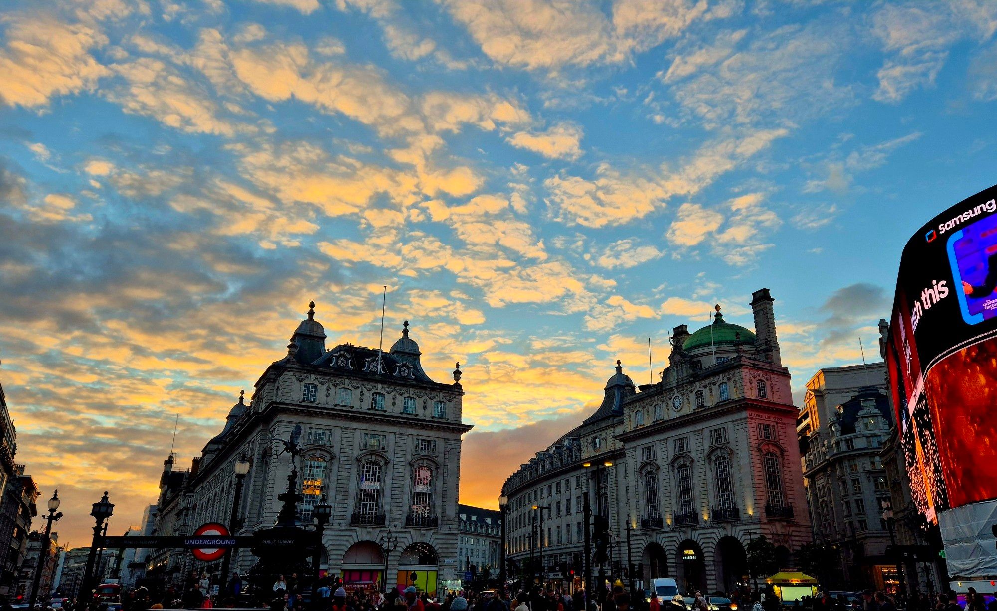 Piccadilly Circus rooftops and neon with a blue sky and orange sunset-filled clouds and an Underground stop in the foreground