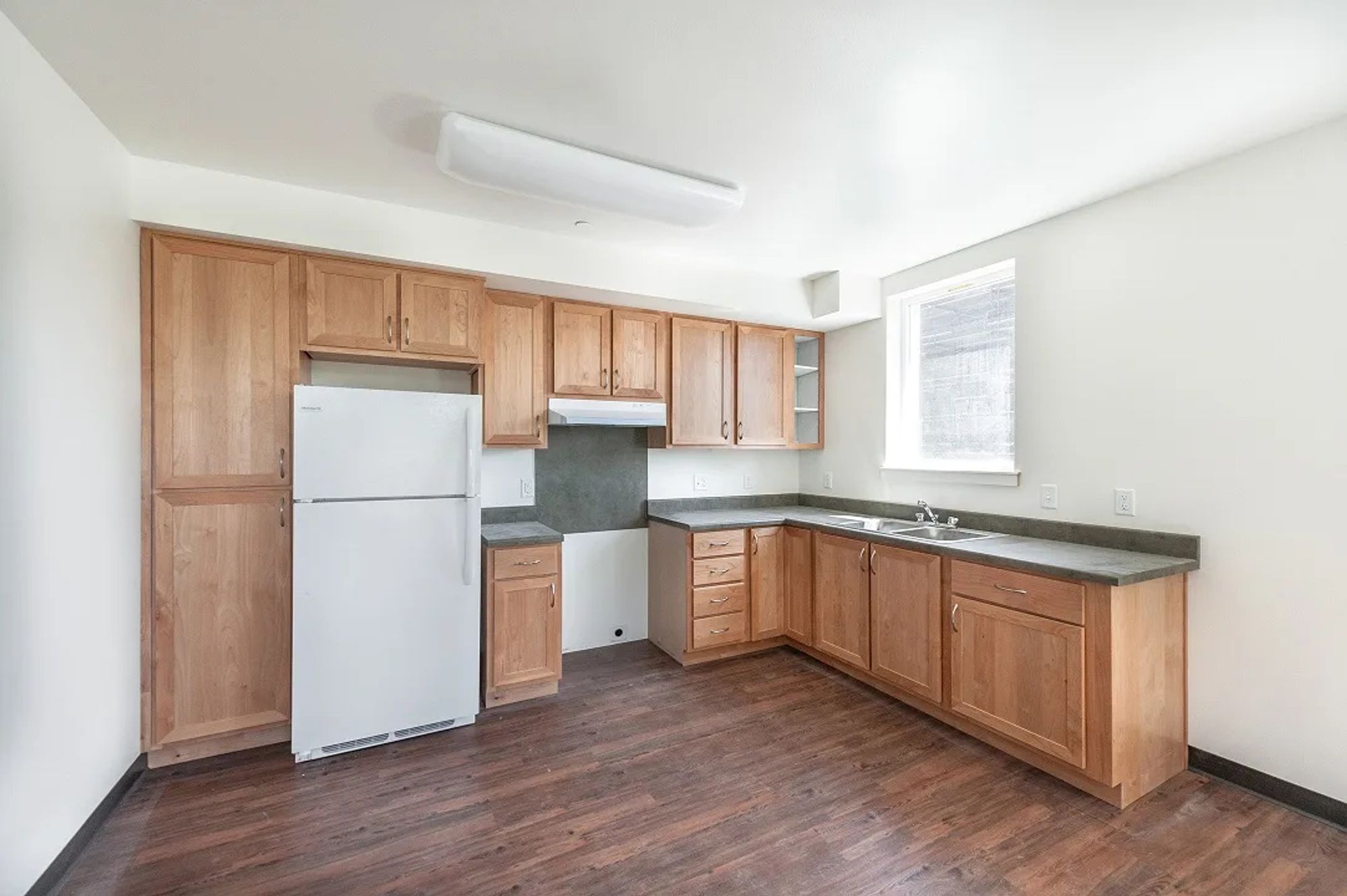 Photo of accessible kitchen. The space where the stove would be is empty in this photo, but a fridge and lots of cabinets sit against the walls in an L shape. The room has fake wood flooring and white walls. Again, there is no way to pull up to the sink, nor any open space beneath it. There is a window above it in the bare, white wall. It's like they think getting a wheelchair IN a room is "access." No consideration for the Disabled person needing to USE the fixtures.