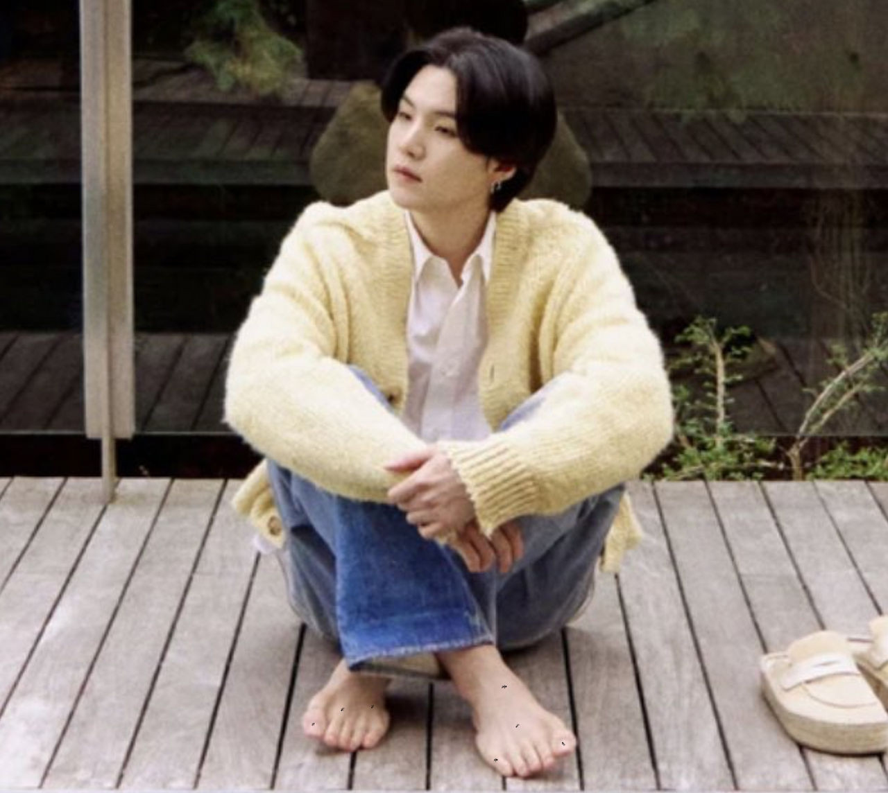 Suga of BTS sitting cross-legged on a deck in a fuzzy yellow cardigan and jeans, barefoot