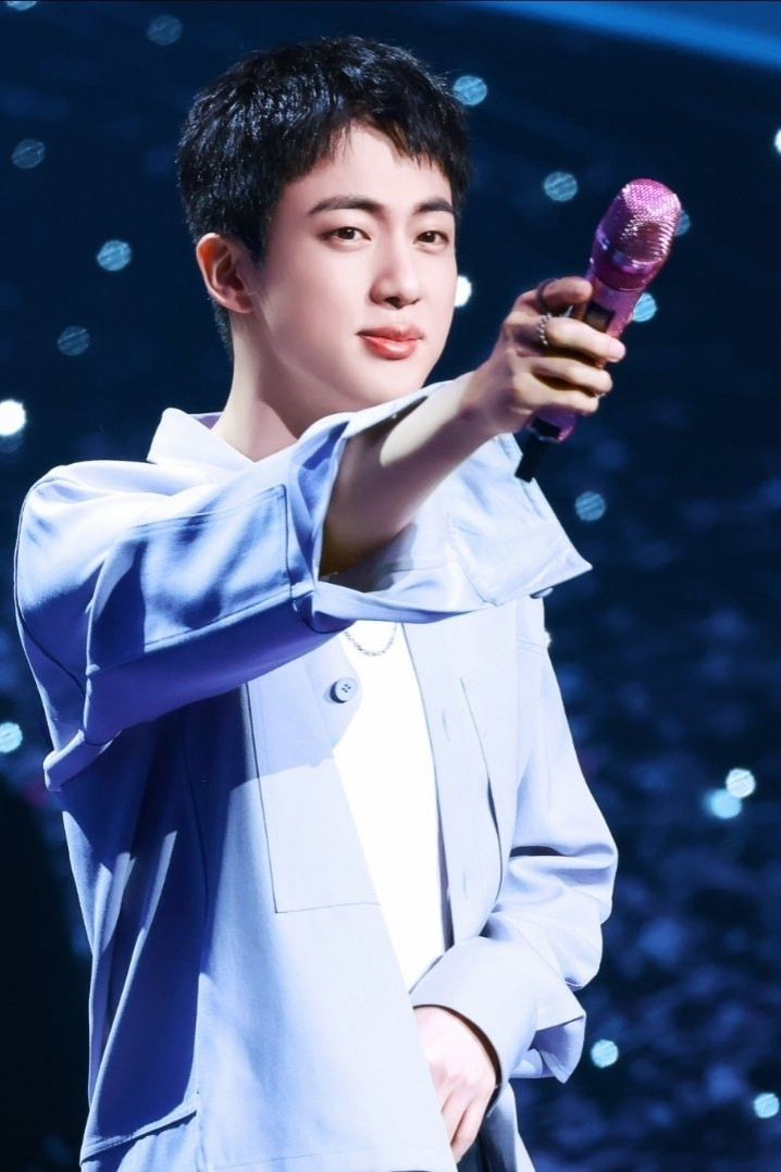 BTS member Jin in a blue shirt smiling and holding out his pink mikrophone to the audience