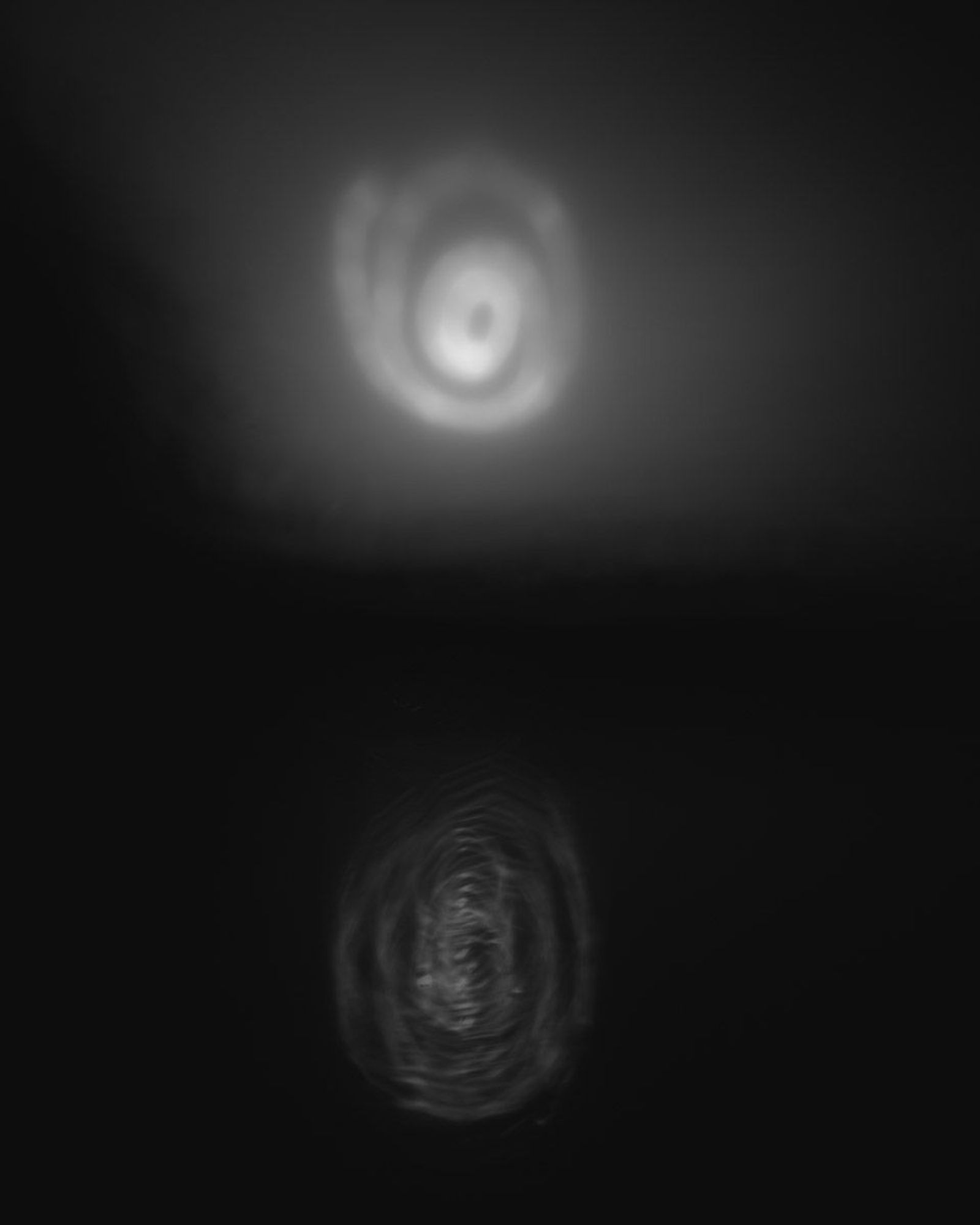 ‘Spiral’ is a black and white abstract landscape photograph from the series ‘dark days irl’. A very dark exposure, it uses intentional camera movement to paint with the sun a rough circling spiral in the sky that’s reflecting shakily and mysteriously below in a rippling body of water. The entire image is dark, blurry and ambiguous with a lake, line of distant trees or hills and clear, graduated sky with sun drawn spiral. 