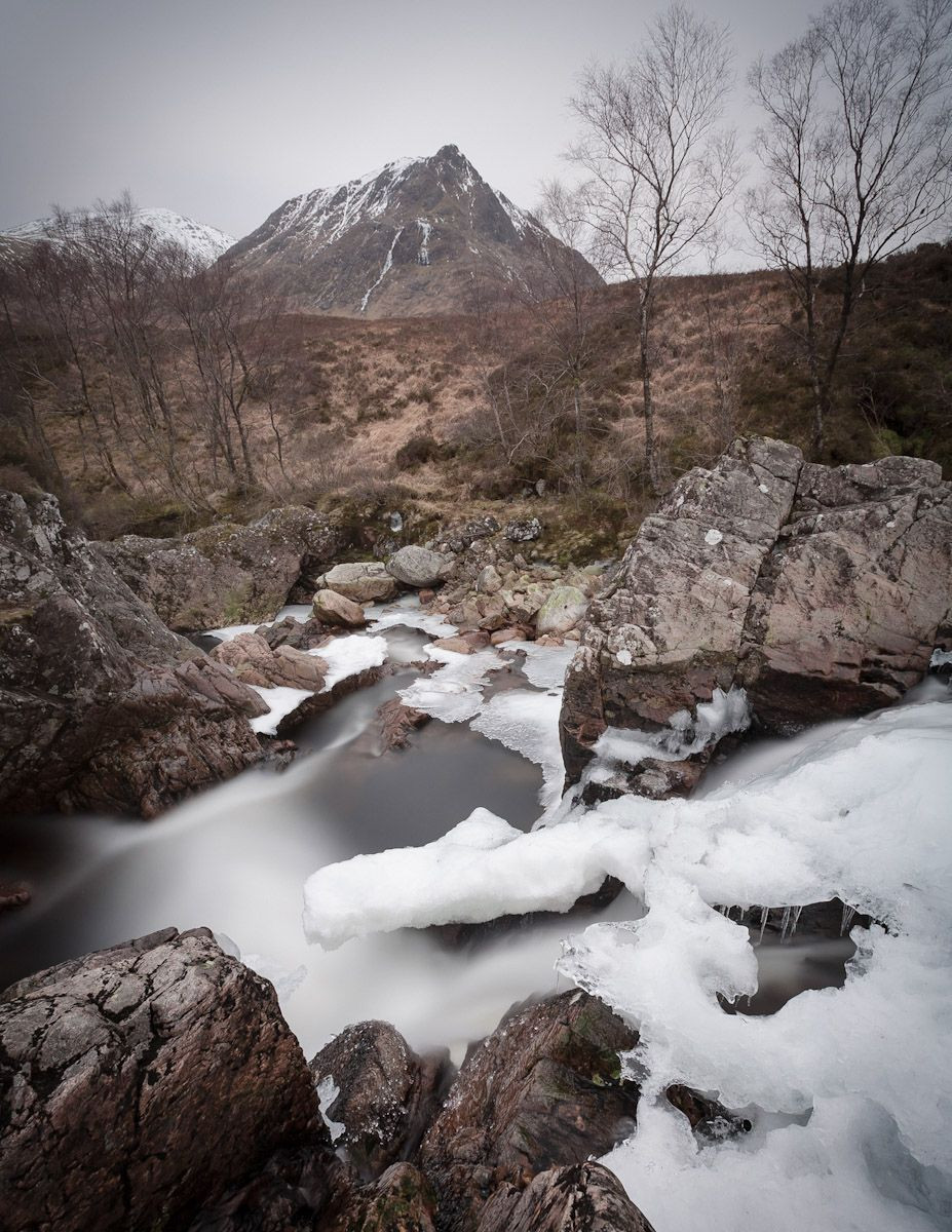 Muted and monochromatic winter colour landscape photograph of a Scottish Highland river tumbling down between giant boulders, it’s banks encased with ice and snow. Spindly trees, a flat grey sky and a rugged, pointed mountain, drab browns and scattered straggly snow. 