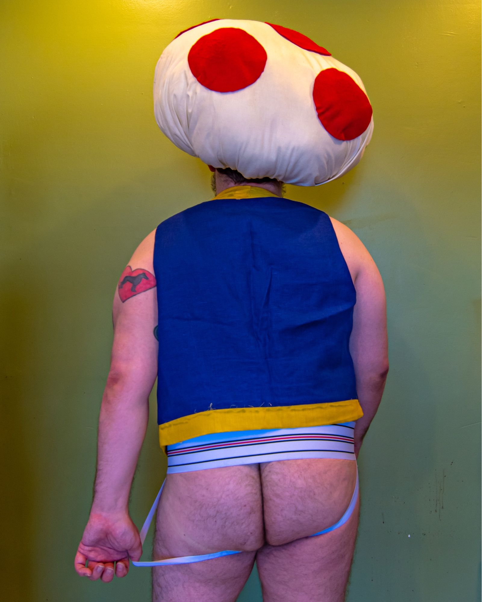 Thicc hairy man, from thighs up and dressed as Toadstool from Super Mario, with a white jockstrap instead of pants, and his left thumb hooked into the strap, pulling it away from his butt.