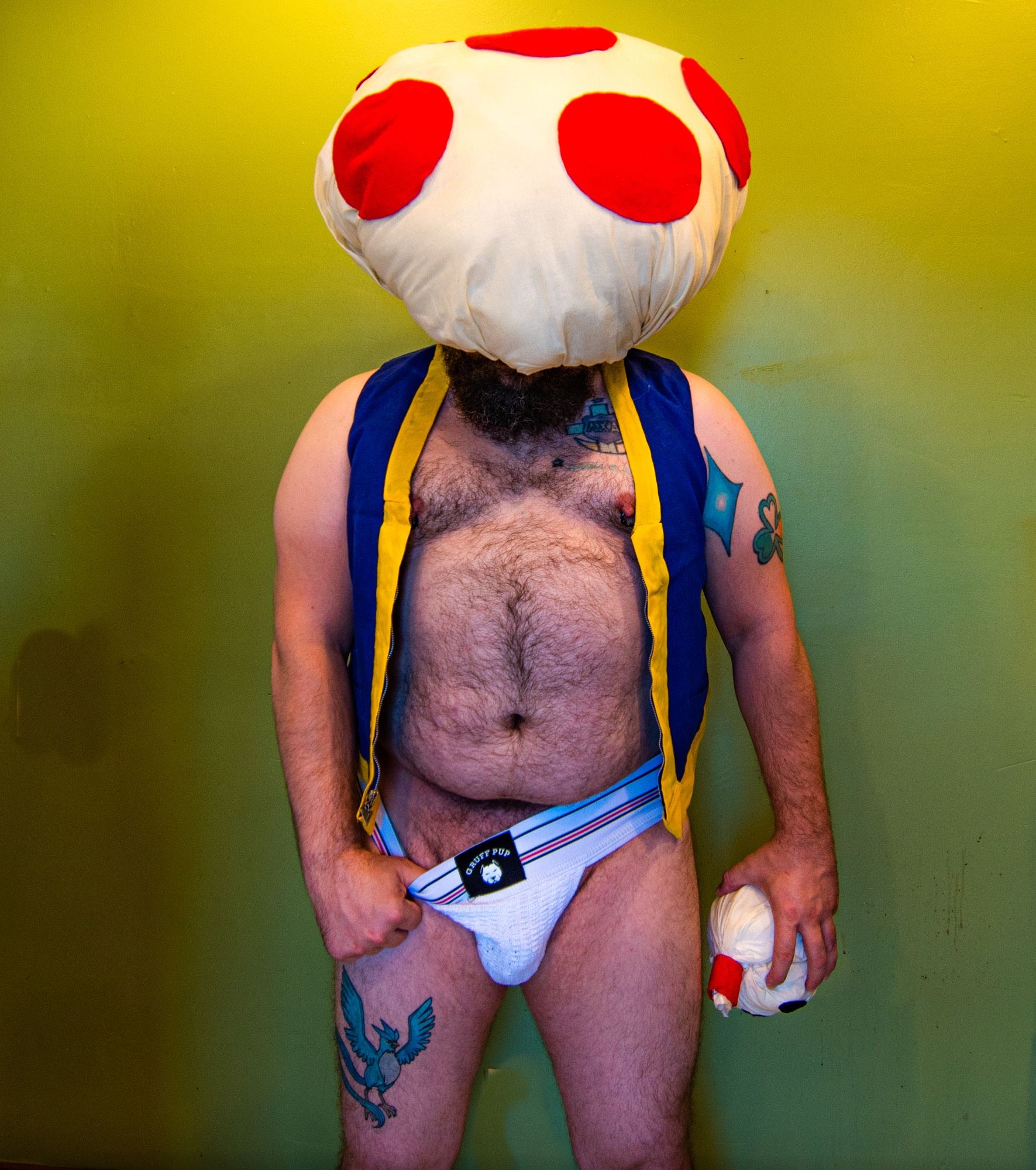 Thicc hairy man, from knees up as Toadstool from Super Mario, with one thumb hooked into the waistband of his jockstrap and his head looking down, with his mushroom headpiece covering his face.