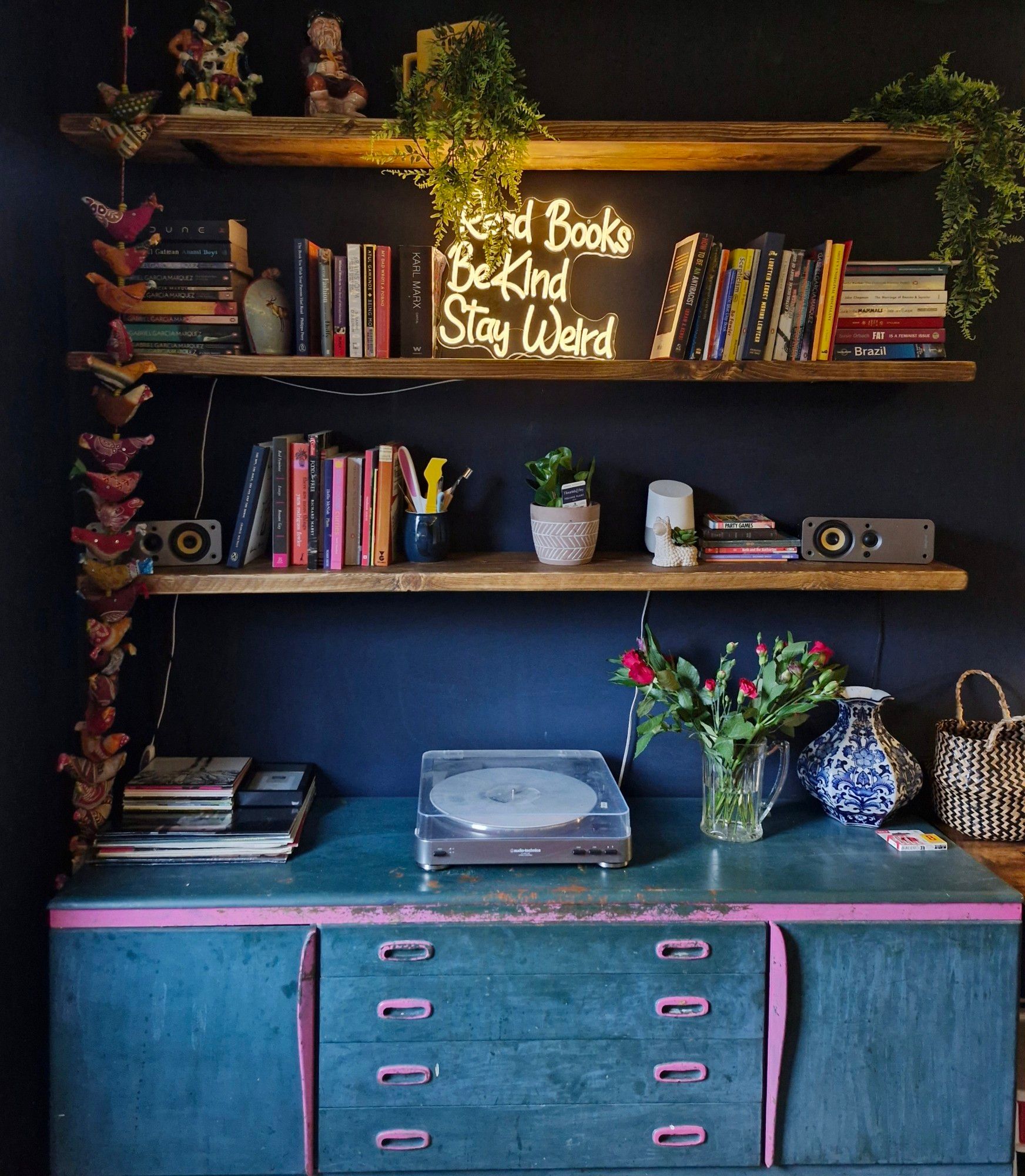 Three bookshelves in a navy room, above a sideboard with record player and vase of flowers. There is a neon light on the middle shelf saying "read books, be kind, stay weird"