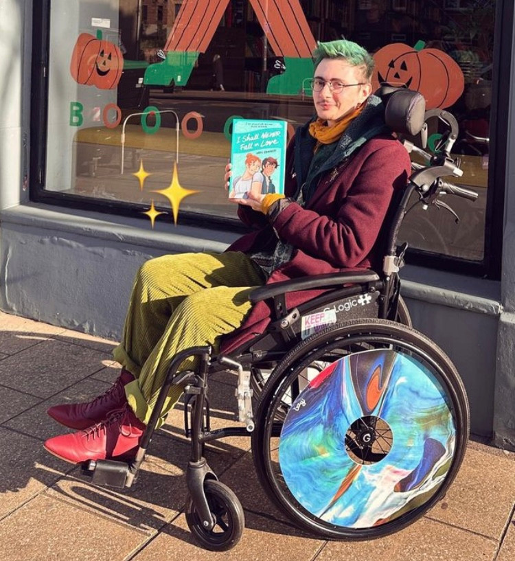 photo of me (white, glasses, green hair and scrappy facial hair, dressed like a hobbit) in the sun in front of a bookshop, holding up my new book