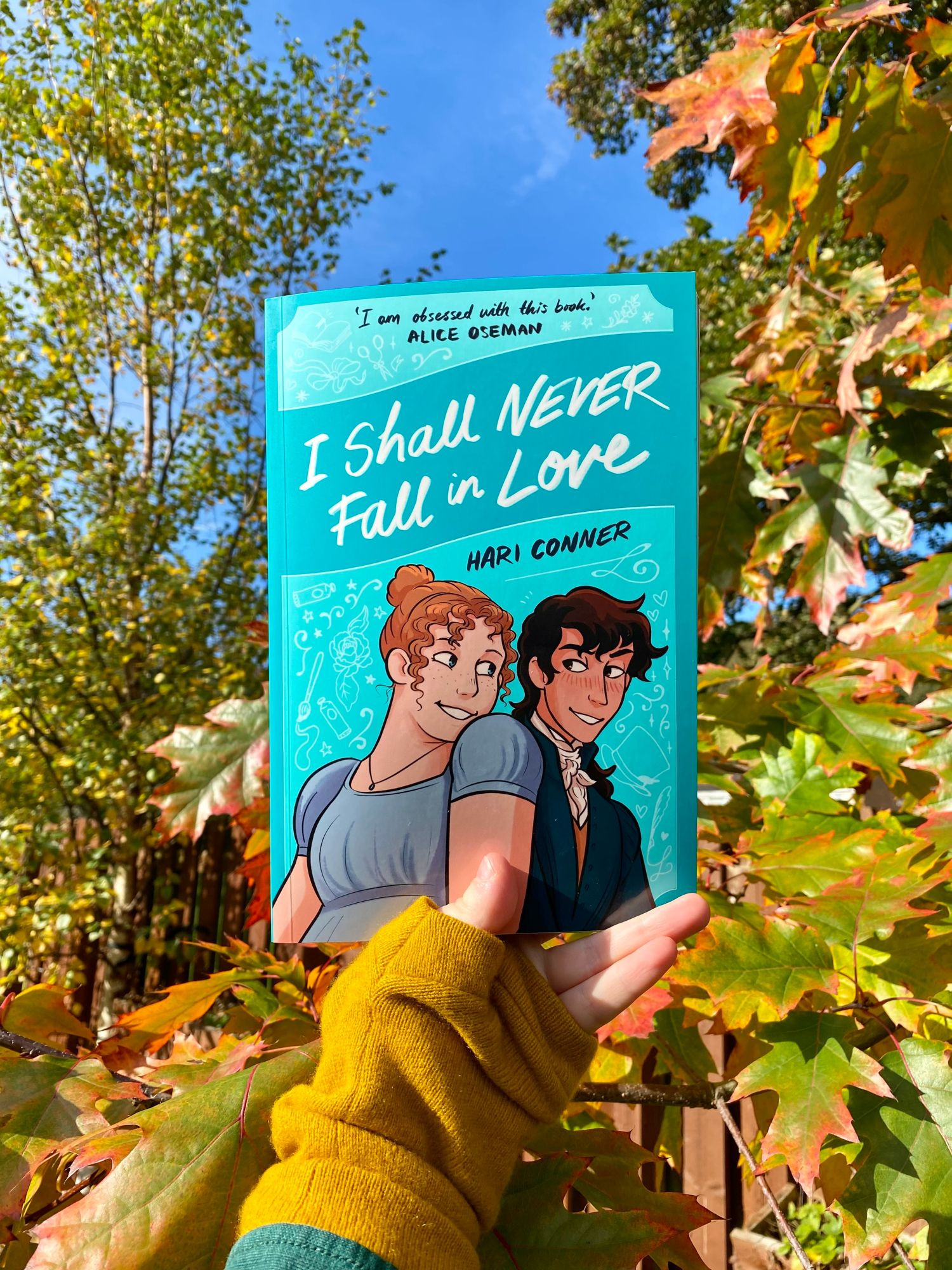 Photo of the book, I Shall Never Fall in Love, held in front of some autumn leaves in the bright sun