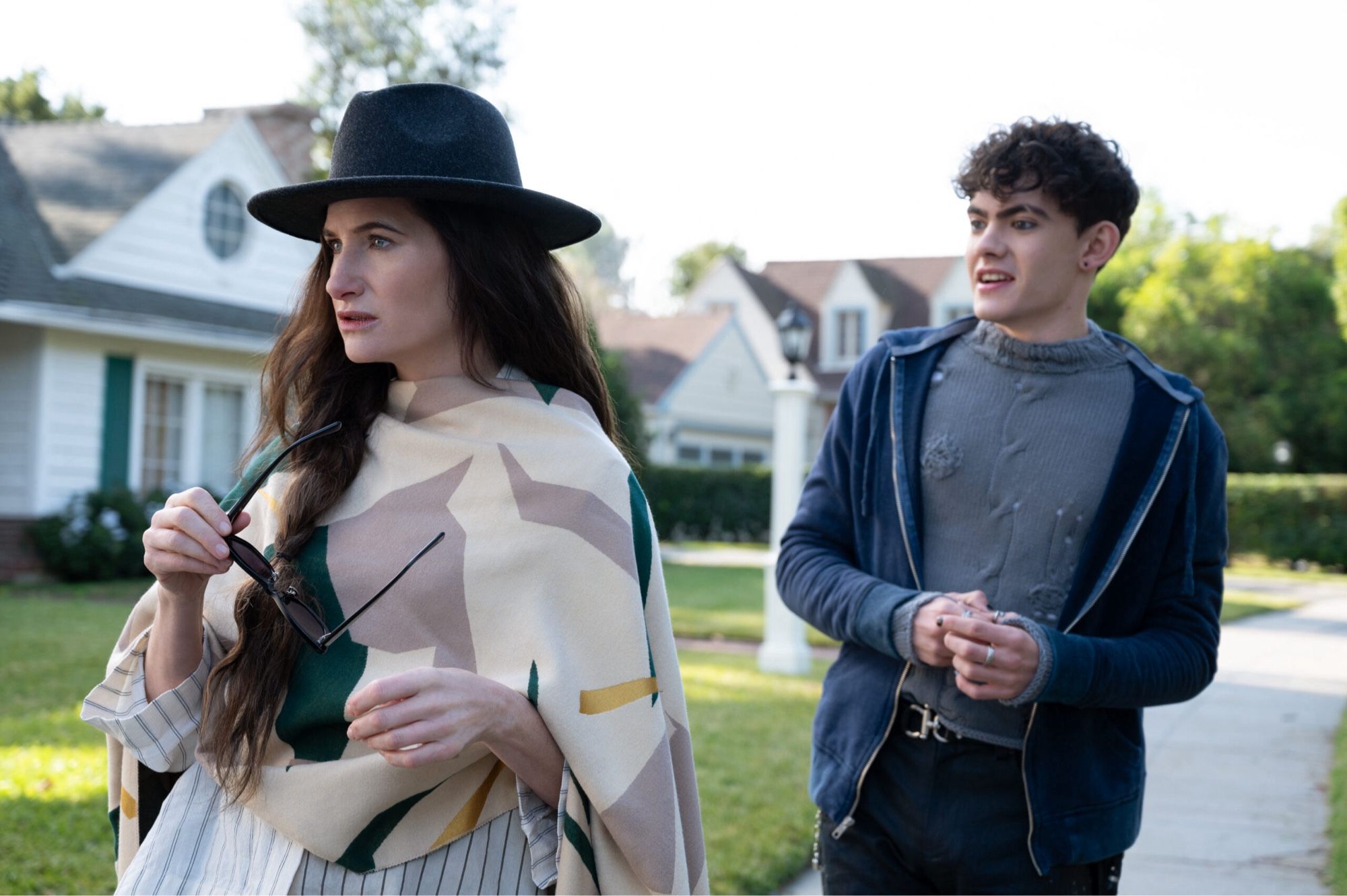 A screenshot of two people. The first, a white woman, looks off to the left of the screen whilst wearing a cream and beige scarf across her torso. The second, a younger male-presenting person in greys and blues, looks at the first person.