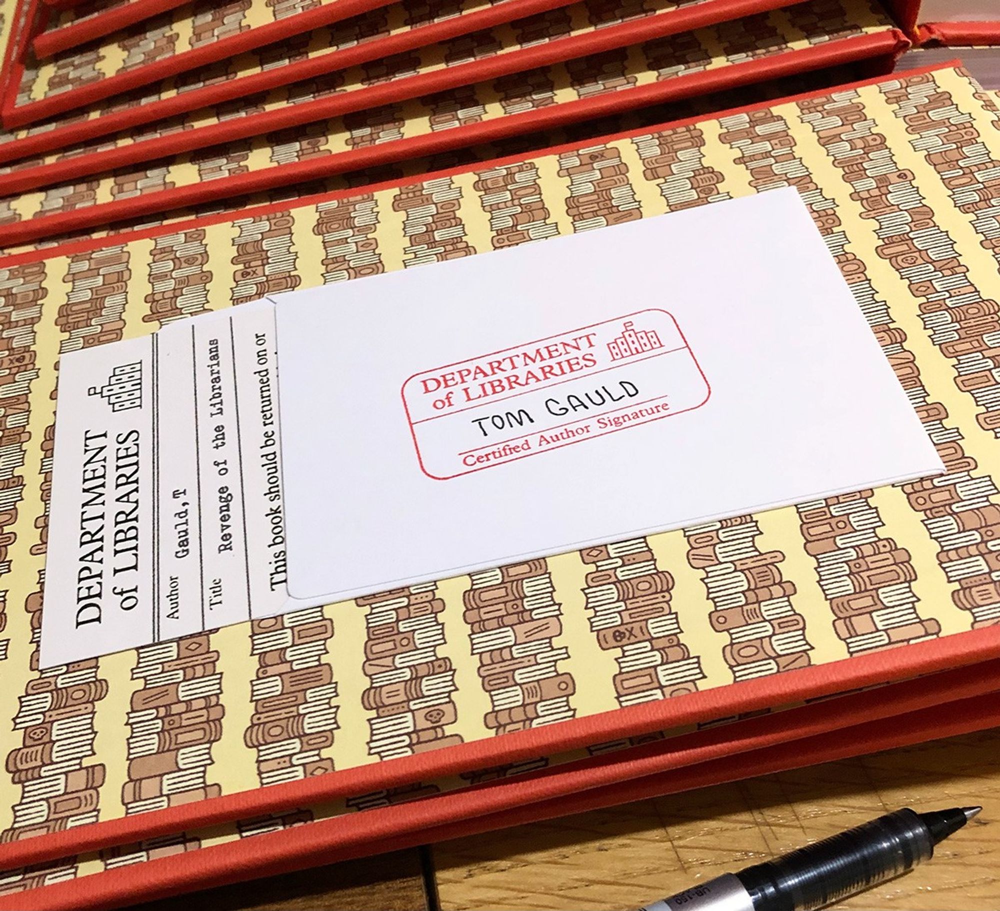 The inside cover of Tom Gauld's book of cartoons 'Revenge of the Librarians' featuring a rubber stamp reading 'Department of Libraries - Certified Author Signature' and Mr Gauld's signature. A pen (uniball eye micro, since you ask) and more copies of the book are also visible.