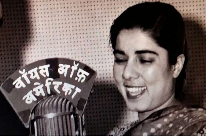 Rajni Kaul as young woman smiling and speaking into old style micropone with topper written in Hindi