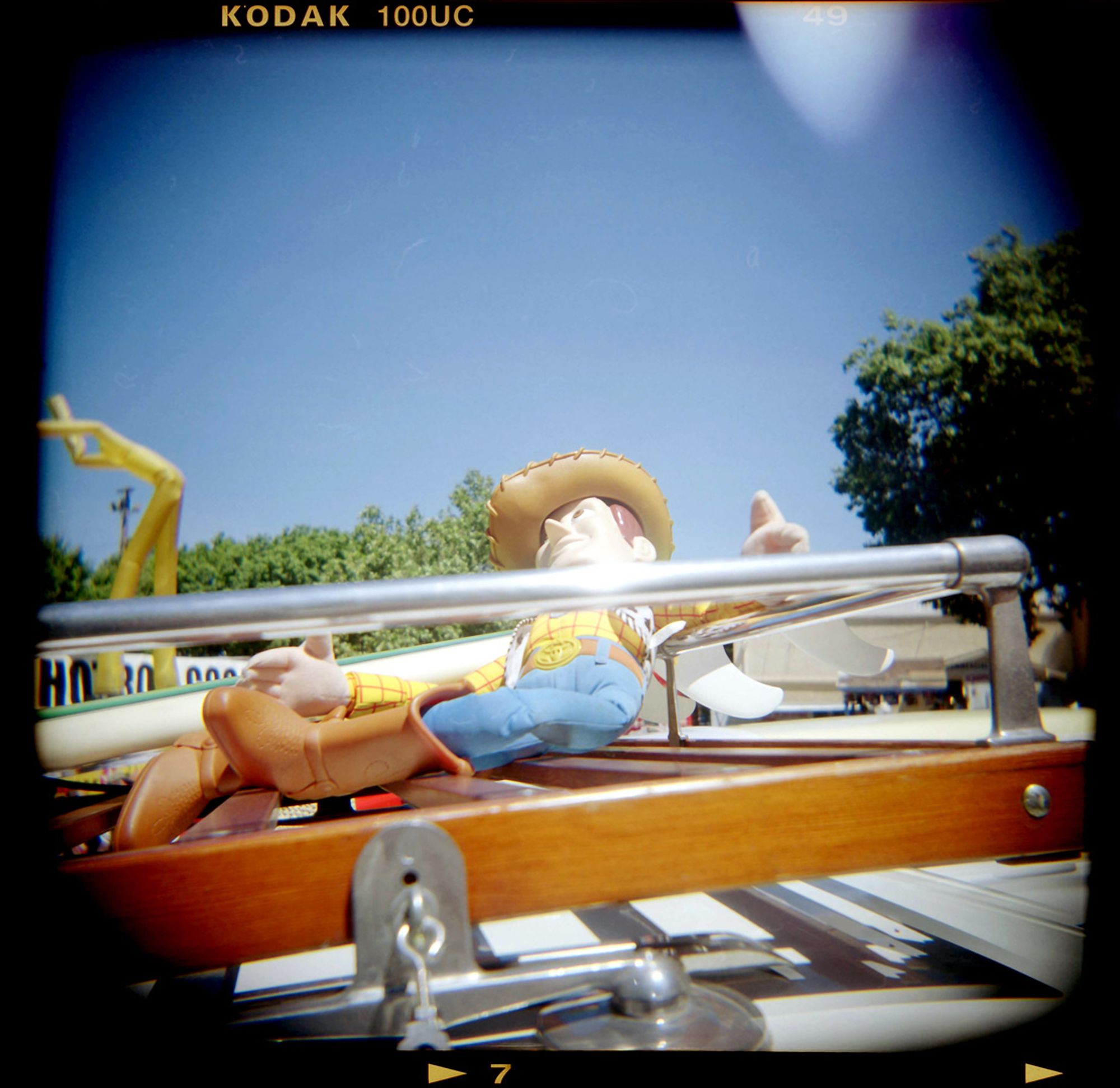 A color photograph showing the roof rack of a vintage woody with a Sheriff Woody Pride doll from Toy Story laying back against the rail and a surfboard in the background.