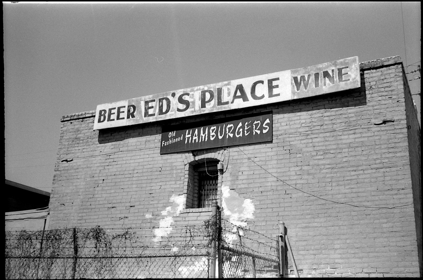 A black and white photograph of the backside of an old brick building with one window and weathered signs for a hamburger, beer and wine joint called Ed's Place in Taylor, Texas.