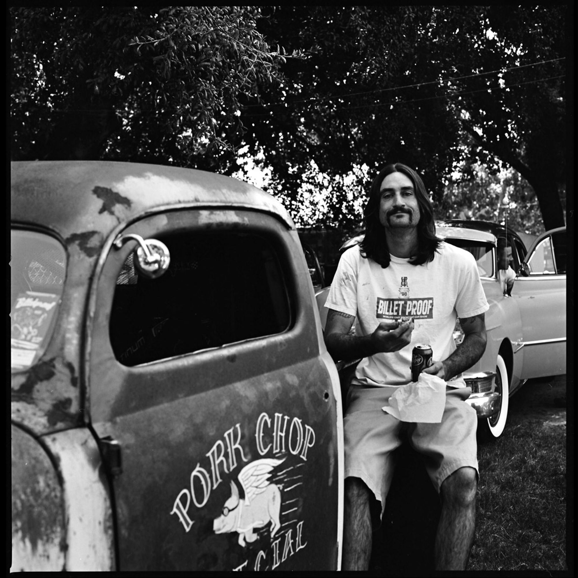 A black and white photograph of a long-haired hot rodder in t-shirt and shorts sitting on the back tire of his car munching on a hotdog and sipping a soda.