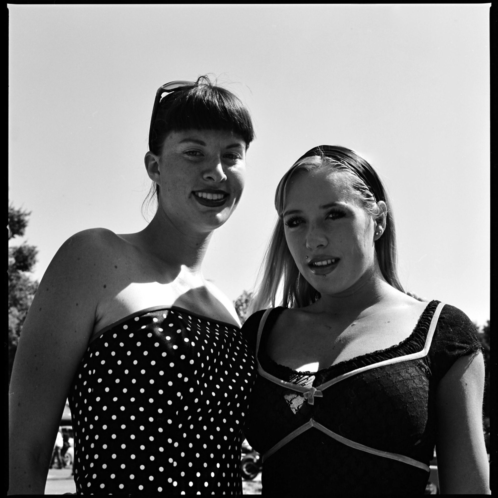 A black and white photograph of two young women at a car show, both are wearing dark colored dresses, one in polka dots and has dark hair and bangs, her sunglasses on top of her head, the other is blonde with a dark hair band. The clear sky is in the background.