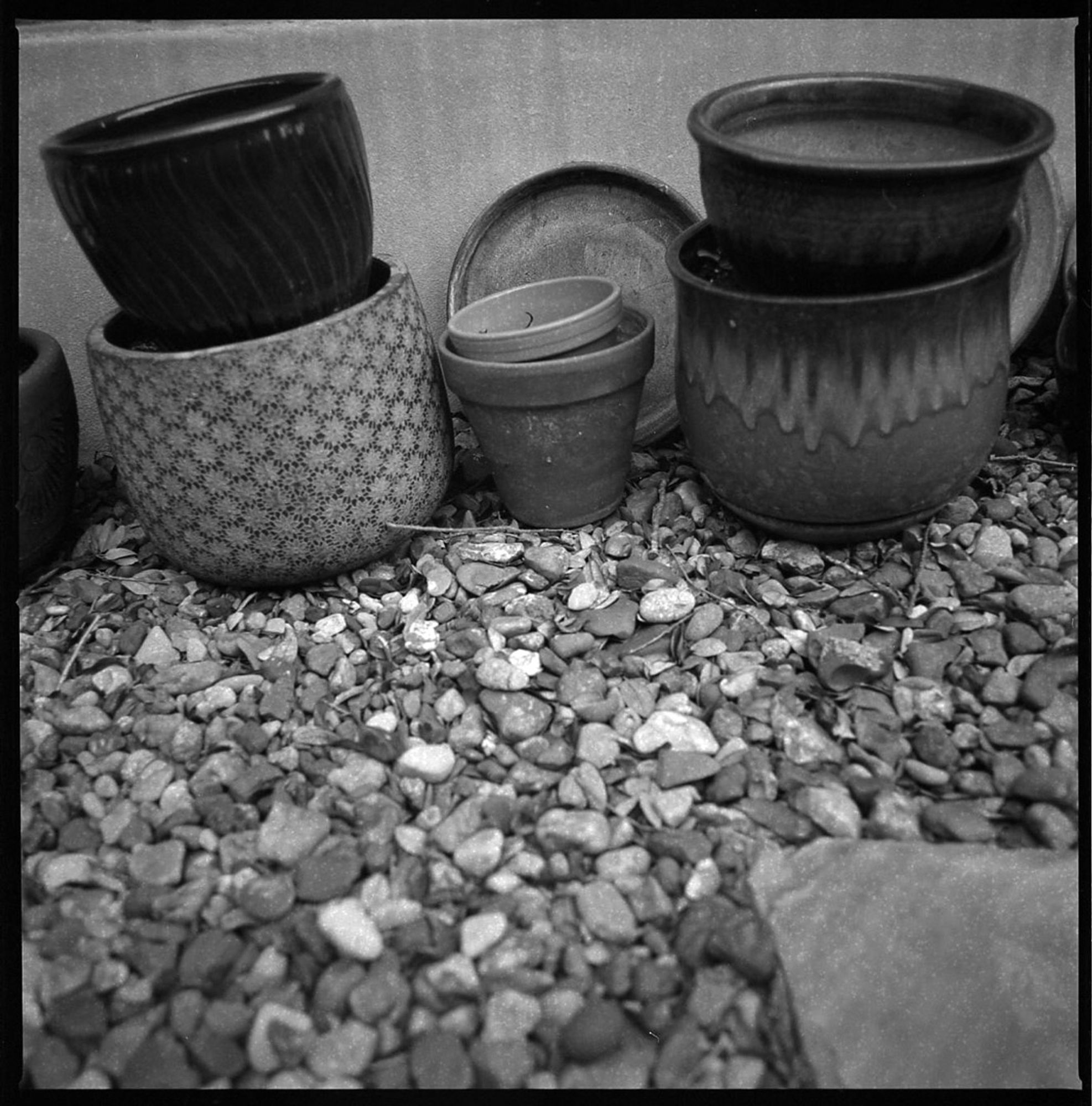 A black and white photograph of stacked garden pots and saucers, up against a concrete foundation, sitting on river rock with a small piece of a flagstone step peeking in from one corner.