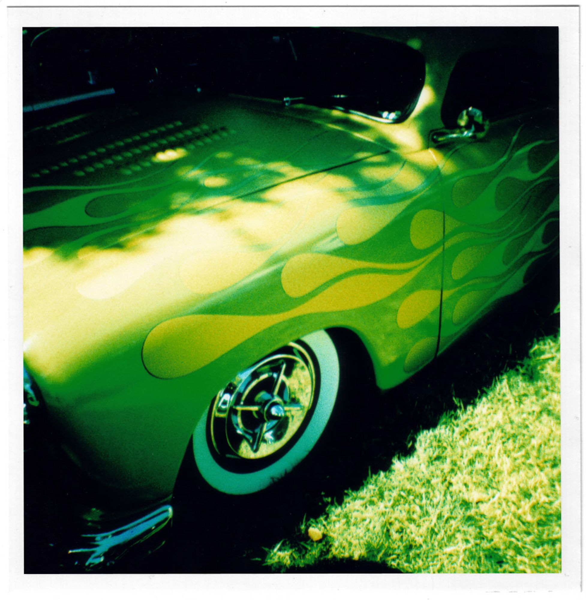 A color photograph of a partially shaded green hotrod's left front end with lighter green flames covering the hood, fender and door, starting out dark at the front of the car in the light and turning light towards the back of the car.