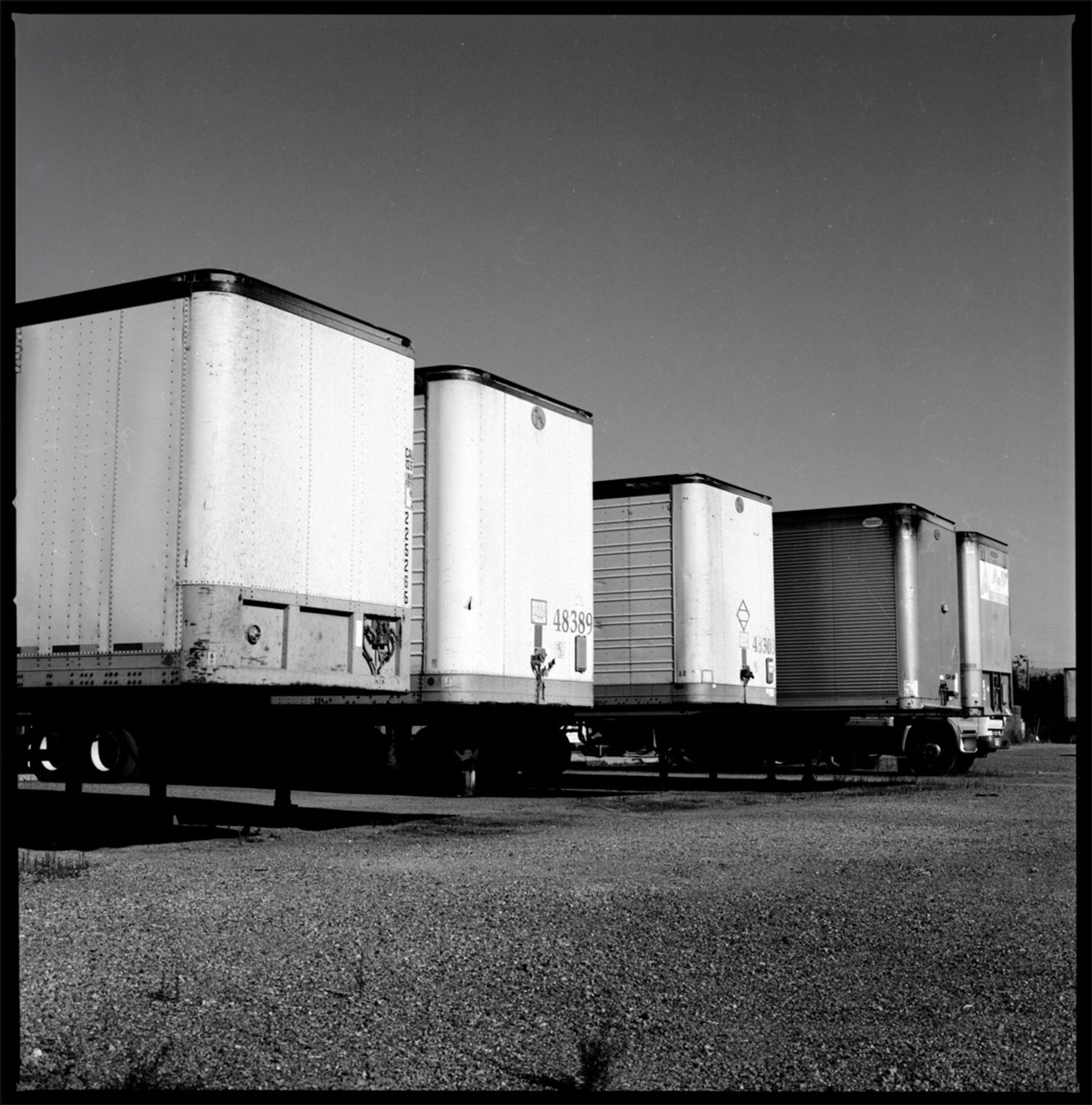 An abstract black and white photograph showing the front ends of five trailers in a row, in two-point perspective, parked on a gravel covered lot.