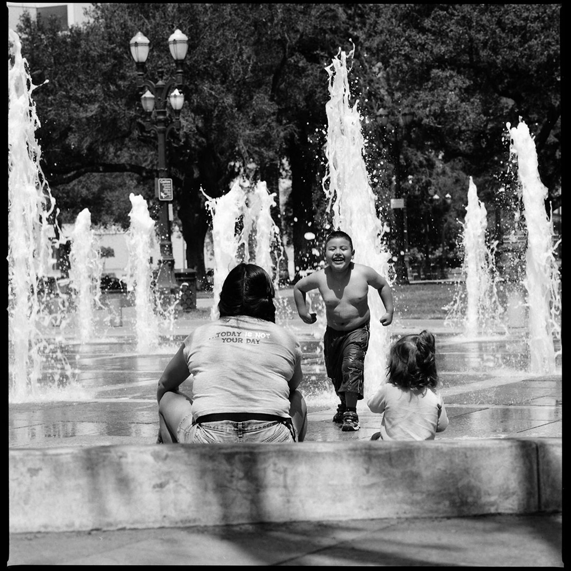 A photograph of a young boy showing off his muscles to his seated mom and little sister while playing in the fountains at Plaza de Cesar Chavez in San Jose, California.