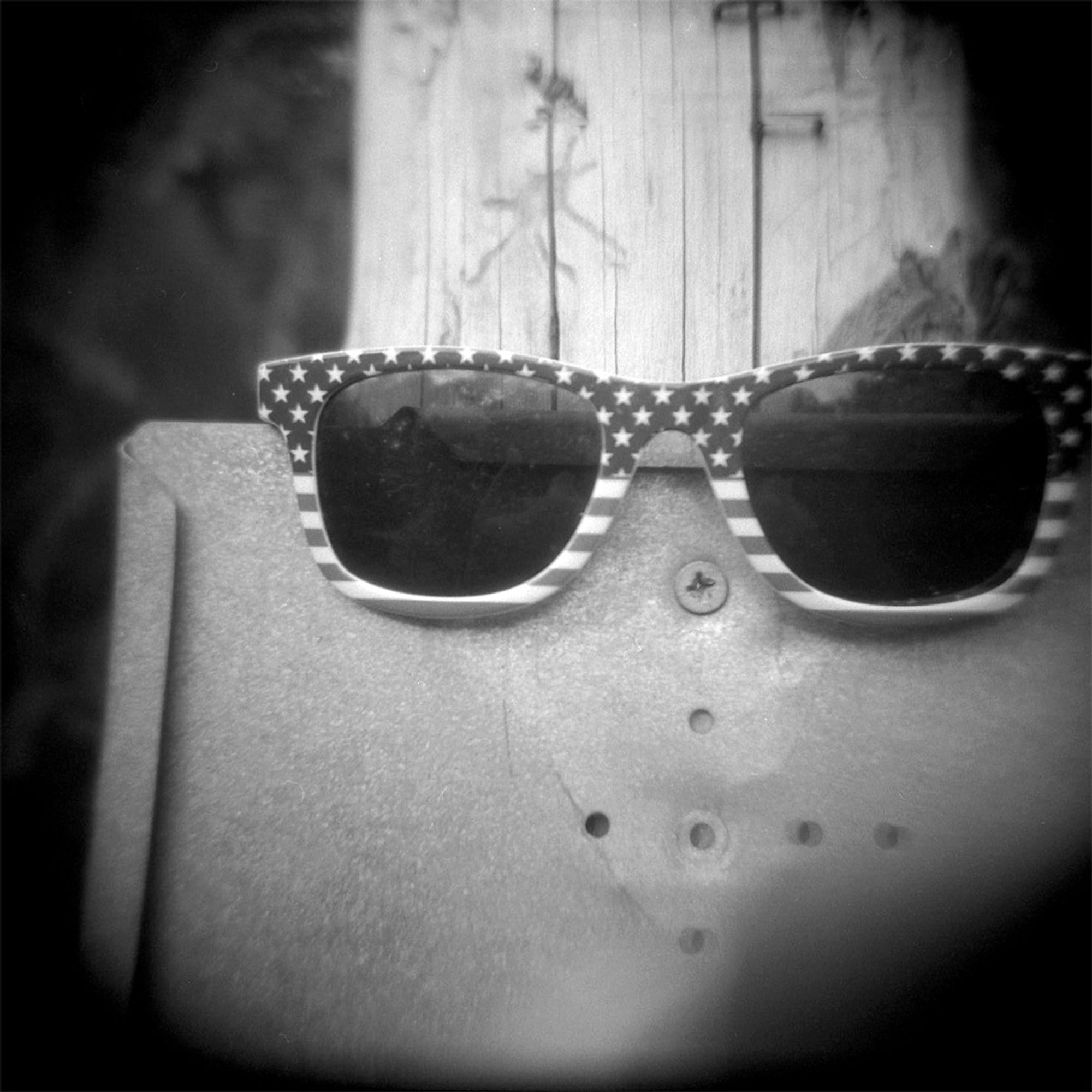 A black and white photograph of what may have been a lost pair of sunglasses, its frame covered in stars and stripes, placed on a sign holder attached to a wooden pole.