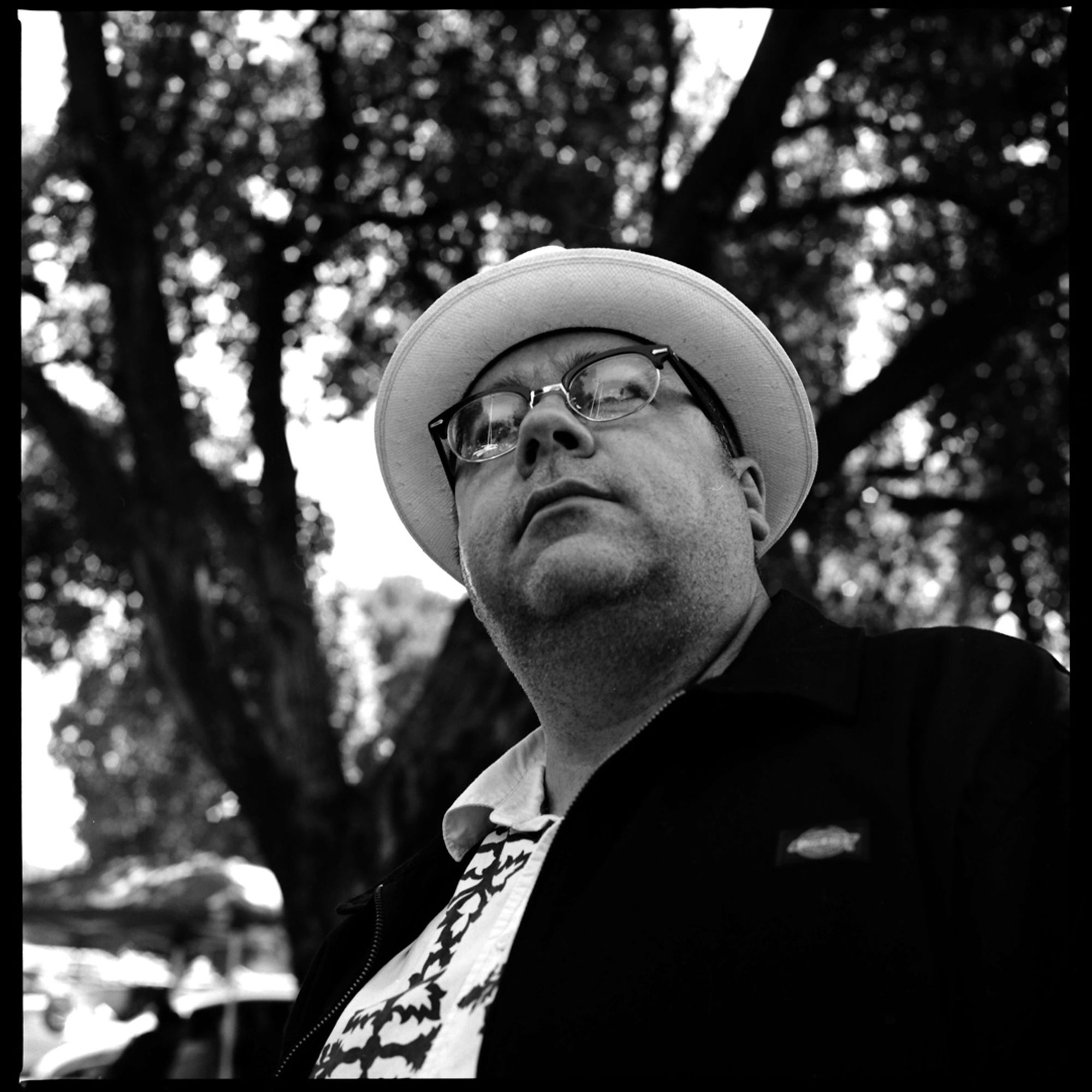 A black and white photograph of a man wearing glasses, a white hat, a fancy white shirt, and dark jacket, looking away from the camera with a canopy of trees in the background.