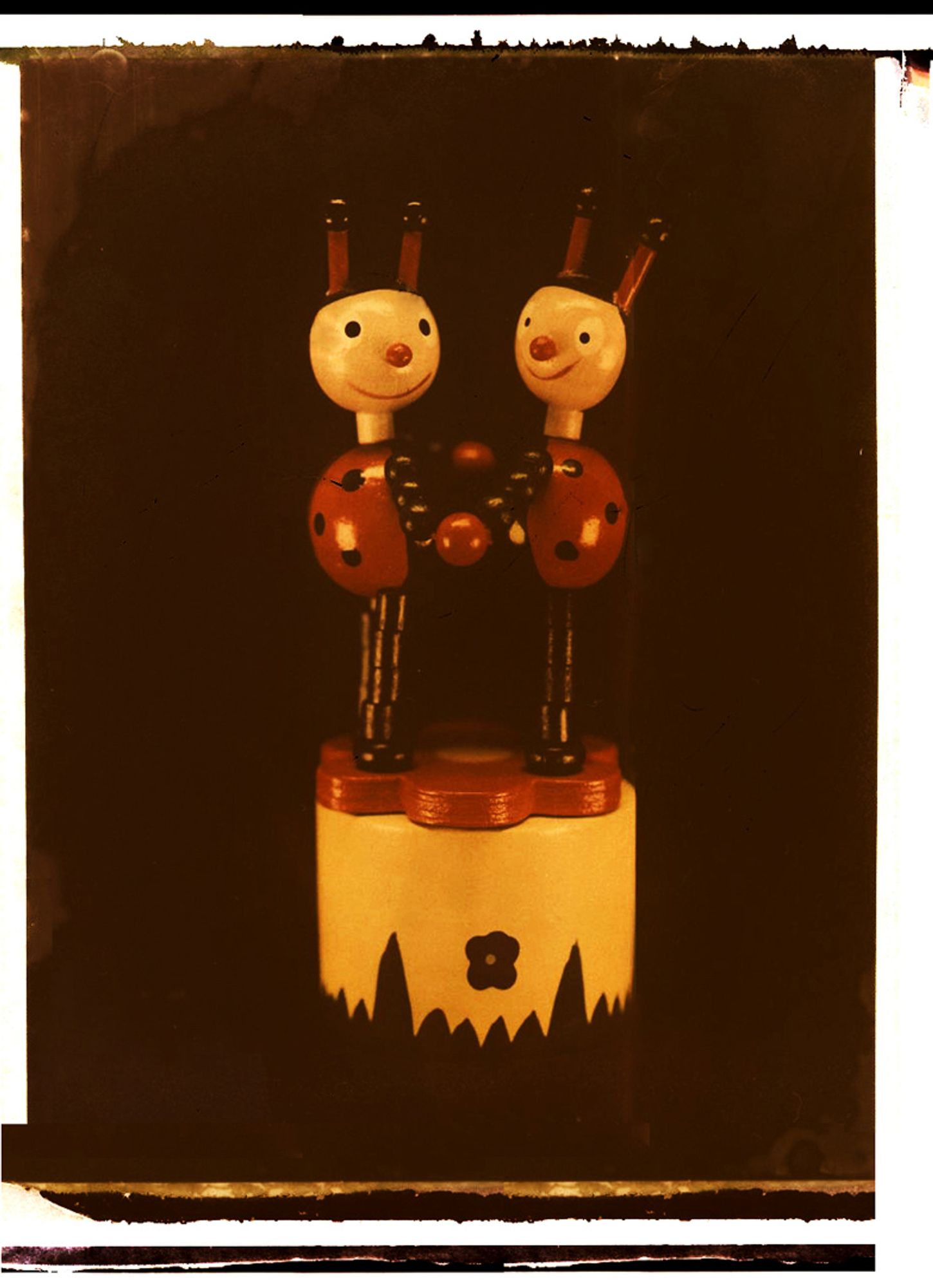 A color instant photograph of a pair of red and black lady bug collapsible push-thumb puppets, holding hands and dancing atop a red flower on a yellow base with green foliage, all set against a black background.