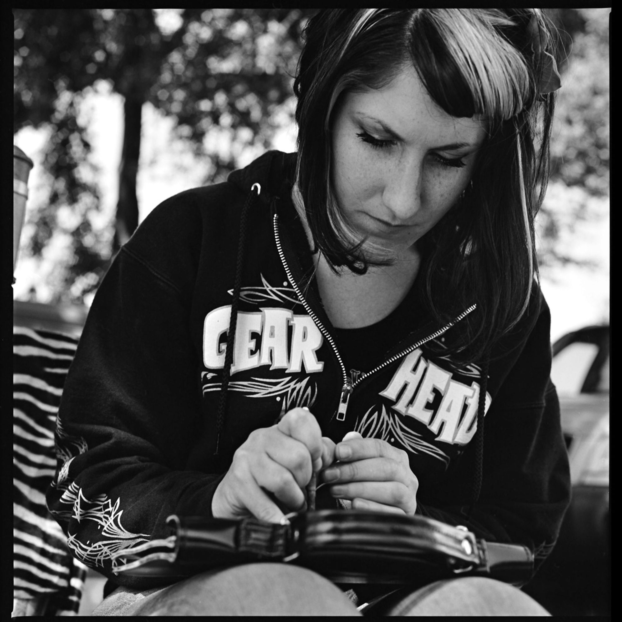 A black and white photograph of a young woman at a car show, sitting while concentrating on pin striping a hand bag, wearing a hoody that has the words gear and head on either side of the zipper.