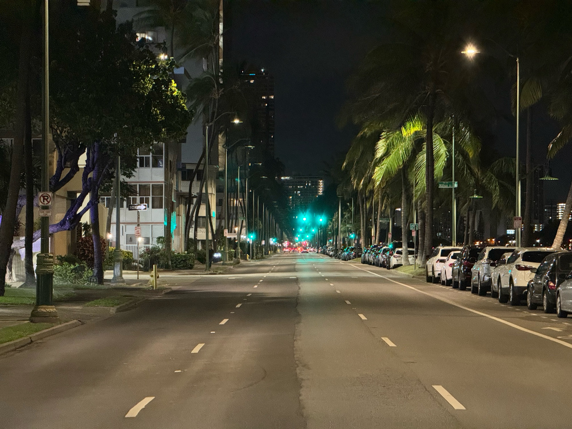 A photo shot down the center of a three lane road at night, with a series of green lights in the distance.