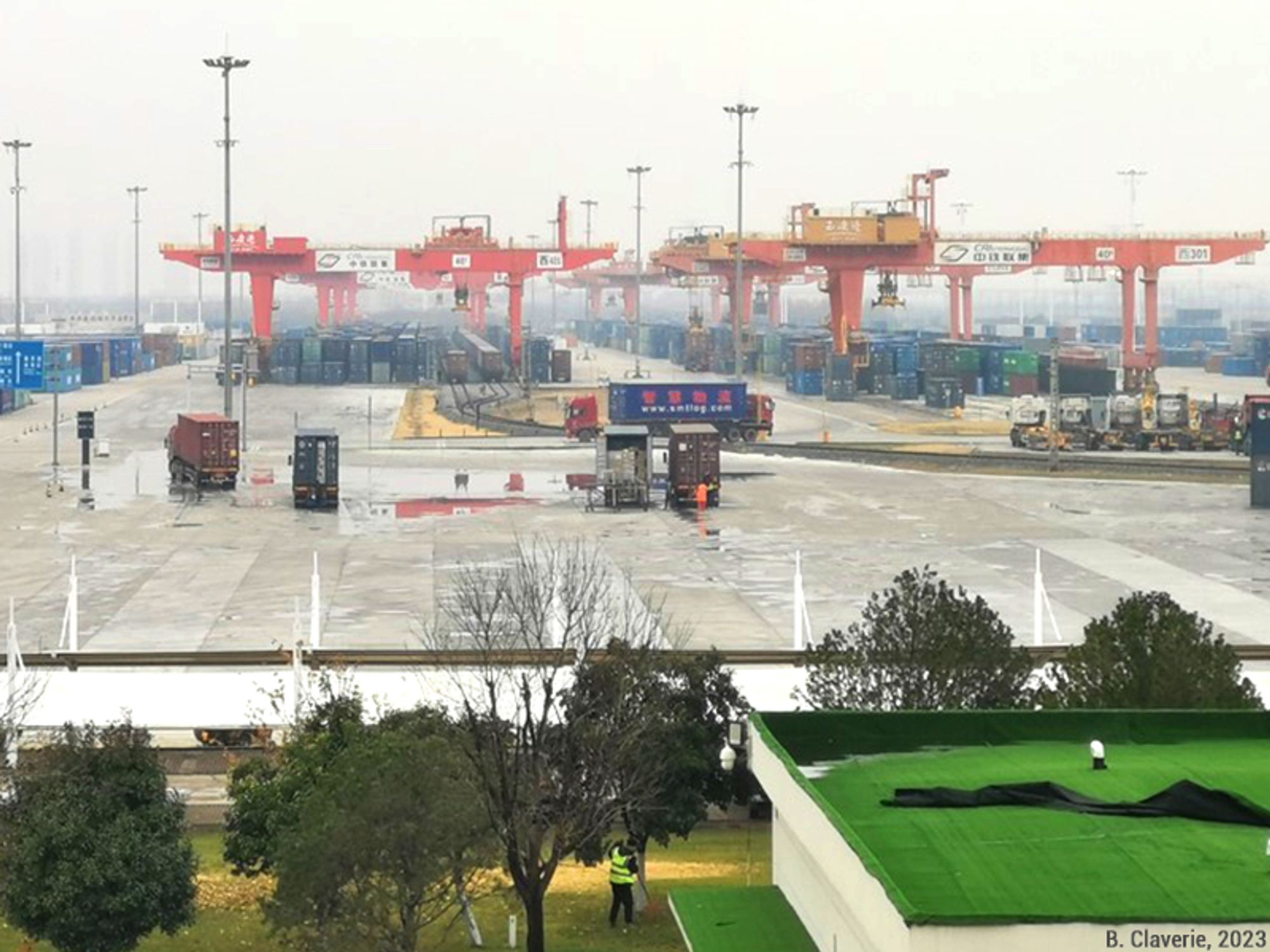 Le tarmac de la plateforme logistique de Xi'an avec des portiques (des grues) rouges, beaucoup de conteneurs entreposés et des camions chargés de conteneurs.