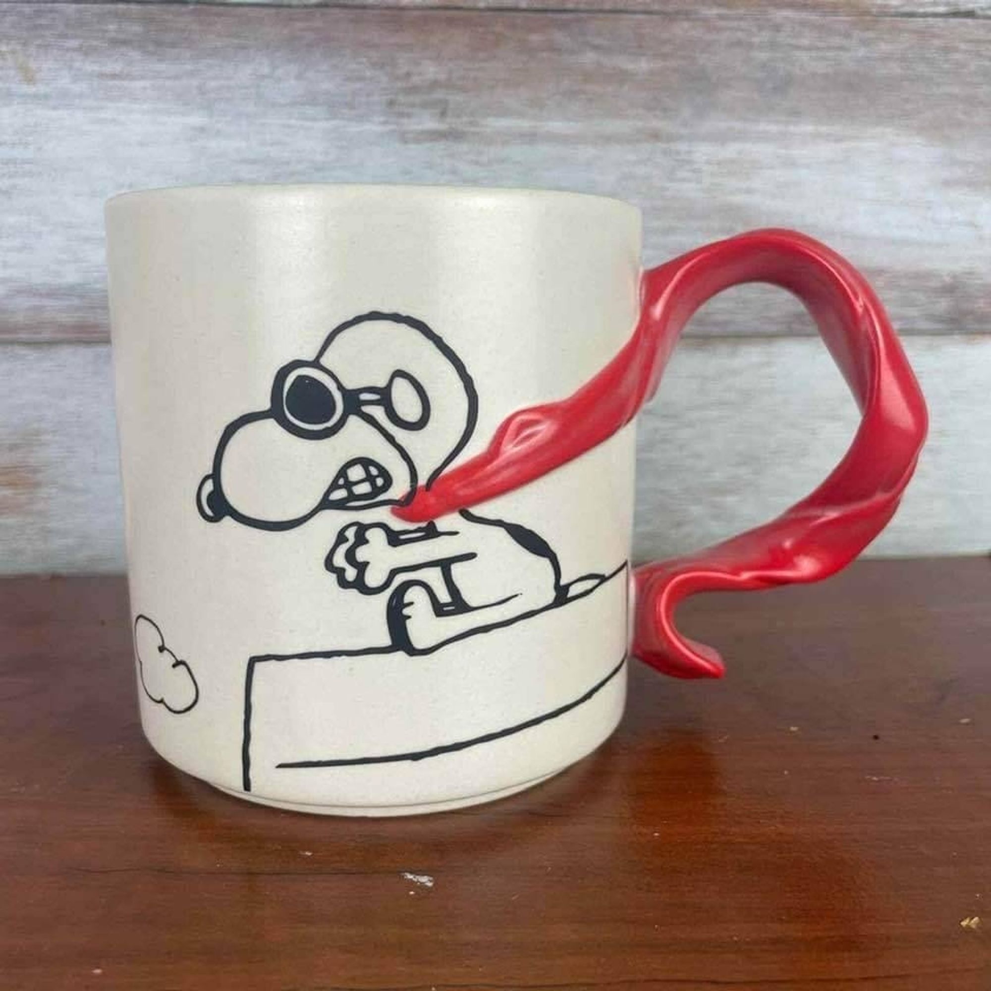 Snoopy est dessiné sur un mug. Il est assis sur sa niche. Il semble conduire une voiture à toute allure. Son écharpe rouge est la anse de la tasse.