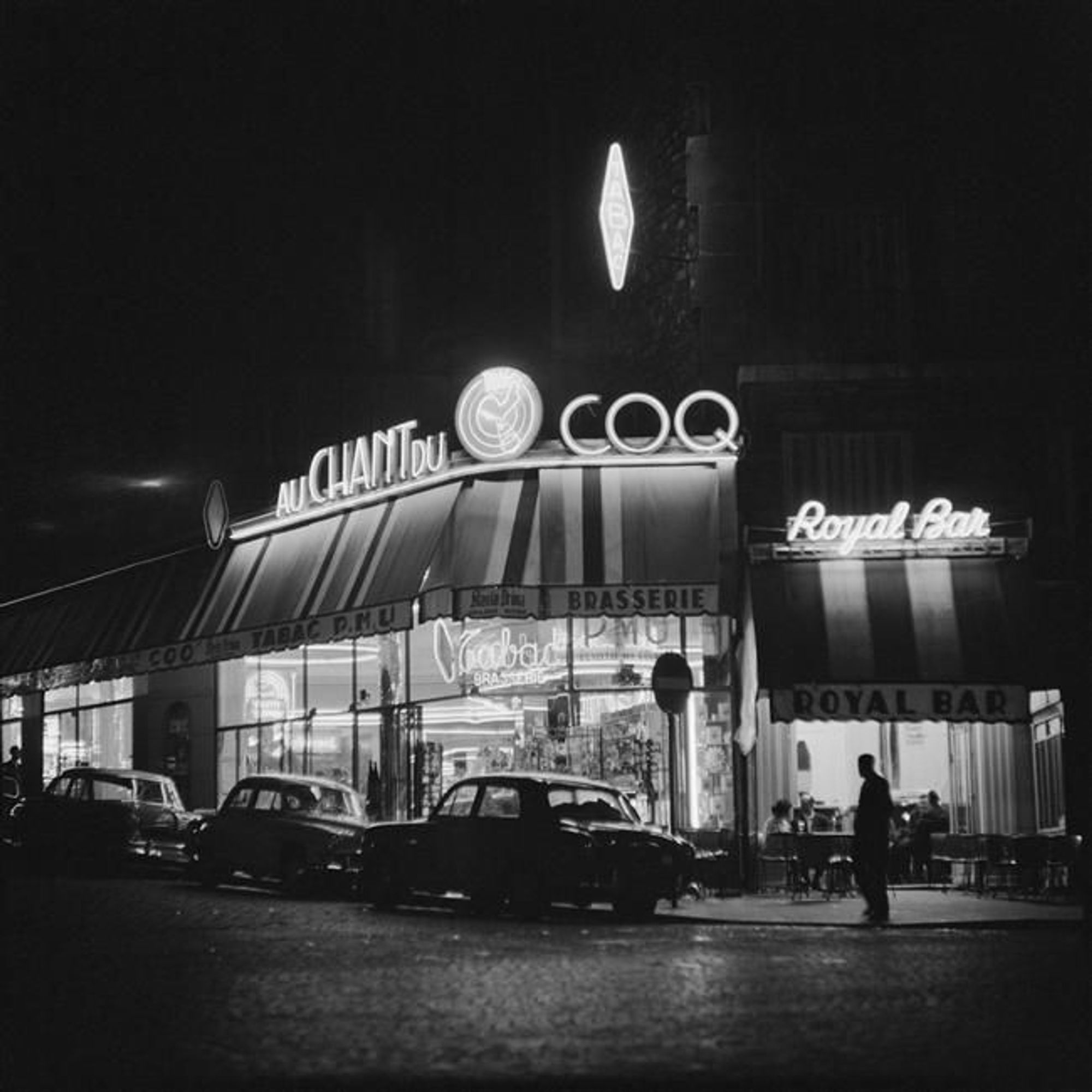 La photo en noir et blanc des années 60 montre une scène de rue nocturne avec une enseigne lumineuse indiquant "AU CHANT DU COQ" sur la façade d'un café brasserie. À droite, on voit une autre enseigne "Royal Bar". Il y a plusieurs voitures garées devant le bâtiment et une silhouette de personne marchant sur le trottoir.