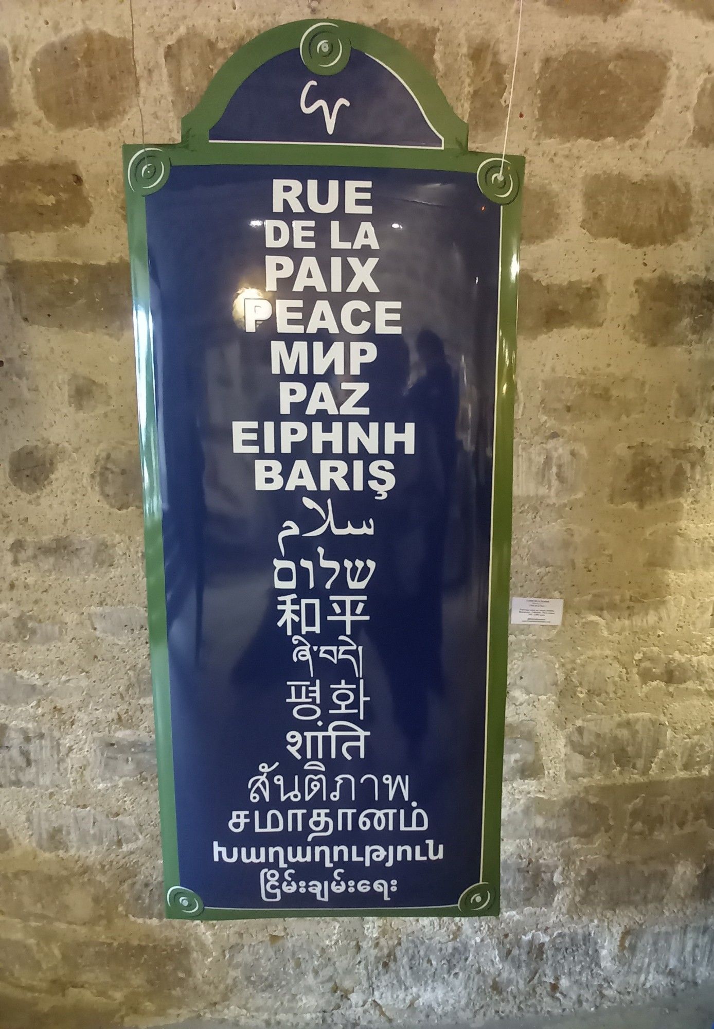 La photo montre un panneau avec l’inscription “RUE DE LA PAIX” en haut, suivi de la traduction du mot “PAIX” dans plusieurs langues différentes. Les traductions sont présentées en colonnes et incluent des langues telles que l’anglais, l’espagnol, le grec, et d’autres écritures non latines comme le chinois, l’hébreu et l’arabe. Le panneau est suspendu sur un mur de pierres apparentes.