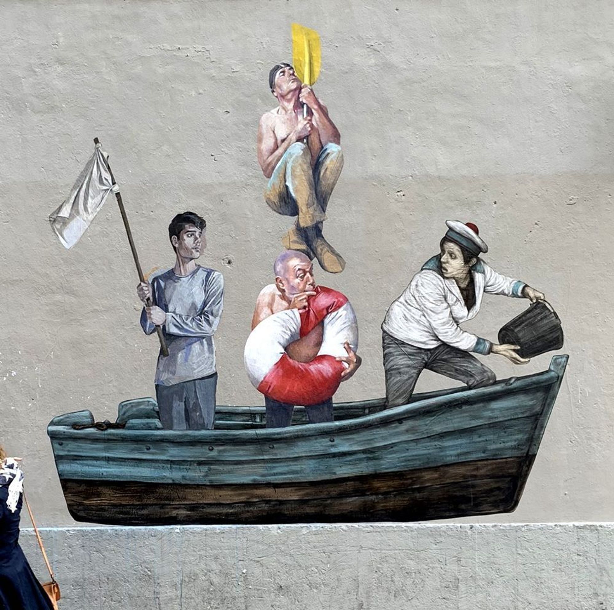 L’image montre une œuvre de street art représentant quatre hommes dans un bateau. L’un agite un drapeau, un autre gonfle une bouée, le troisième vide l’eau du bateau avec un seau, et le dernier est en haut d'une rame verticale.