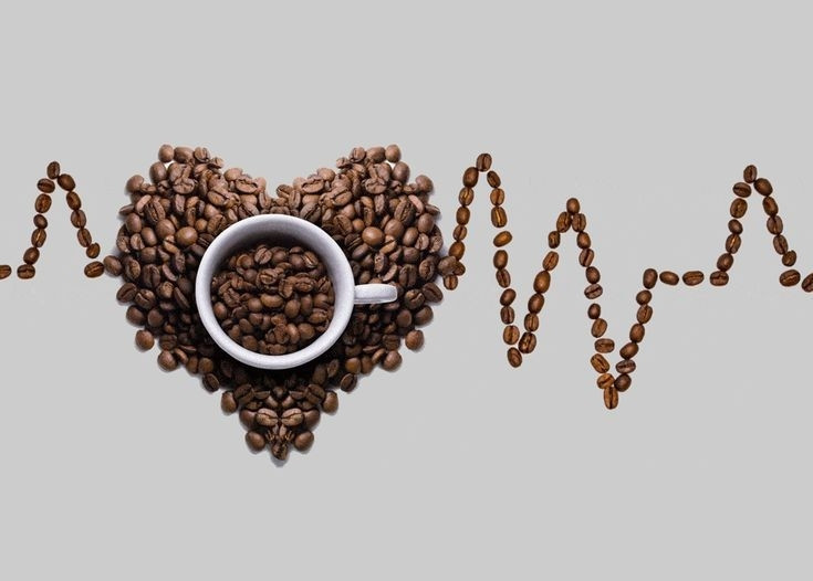 Cette image montre une composition artistique où des grains de café sont disposés pour former un cœur et un électrocardiogramme (ECG). Au centre du cœur, il y a une tasse à café.