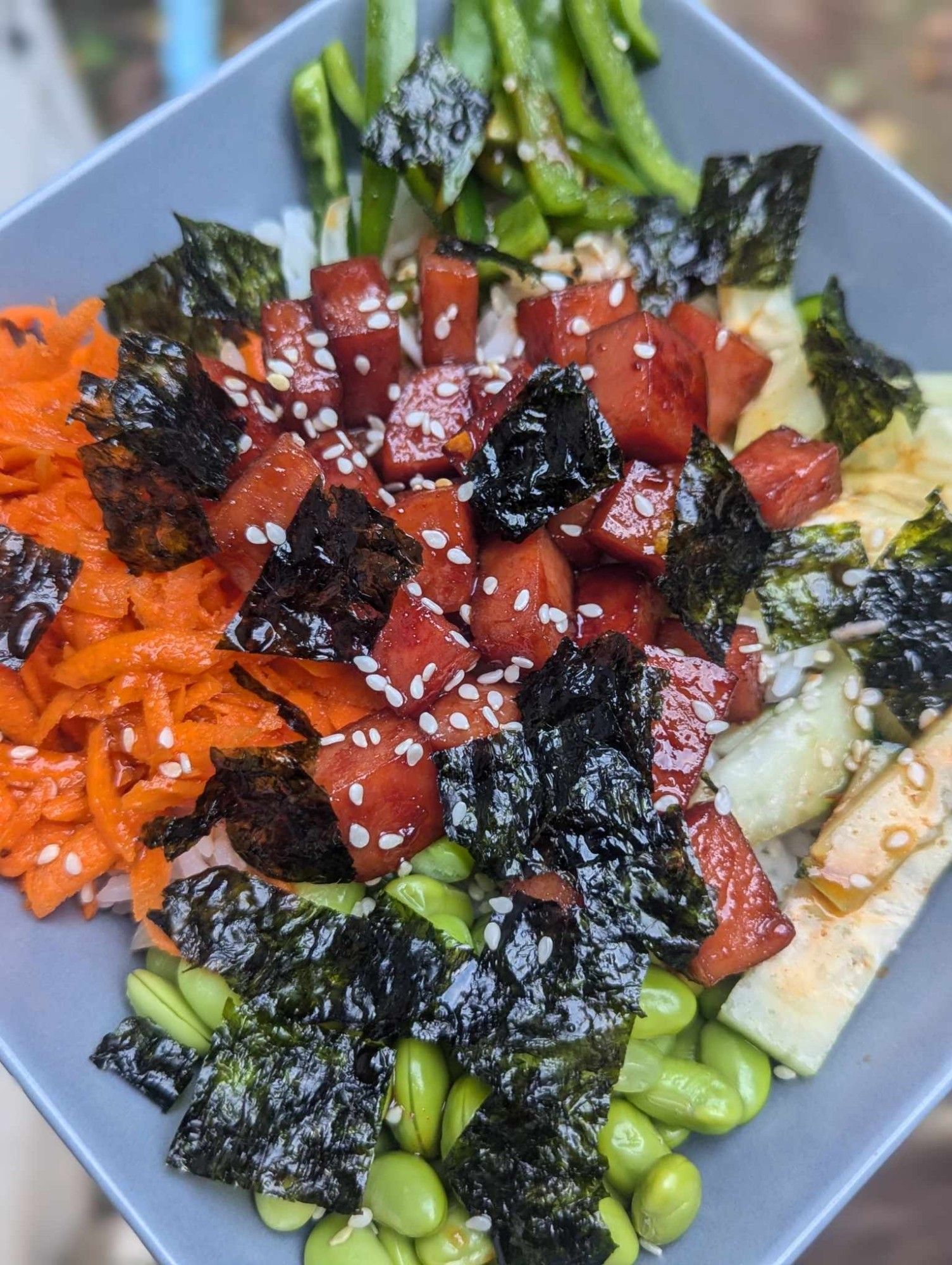 A square grey bowl filled with sushi rice, grated carrot, edamame, cucumber, green pepper, nori, and grilled marinated spam, topped with white sesame seeds.